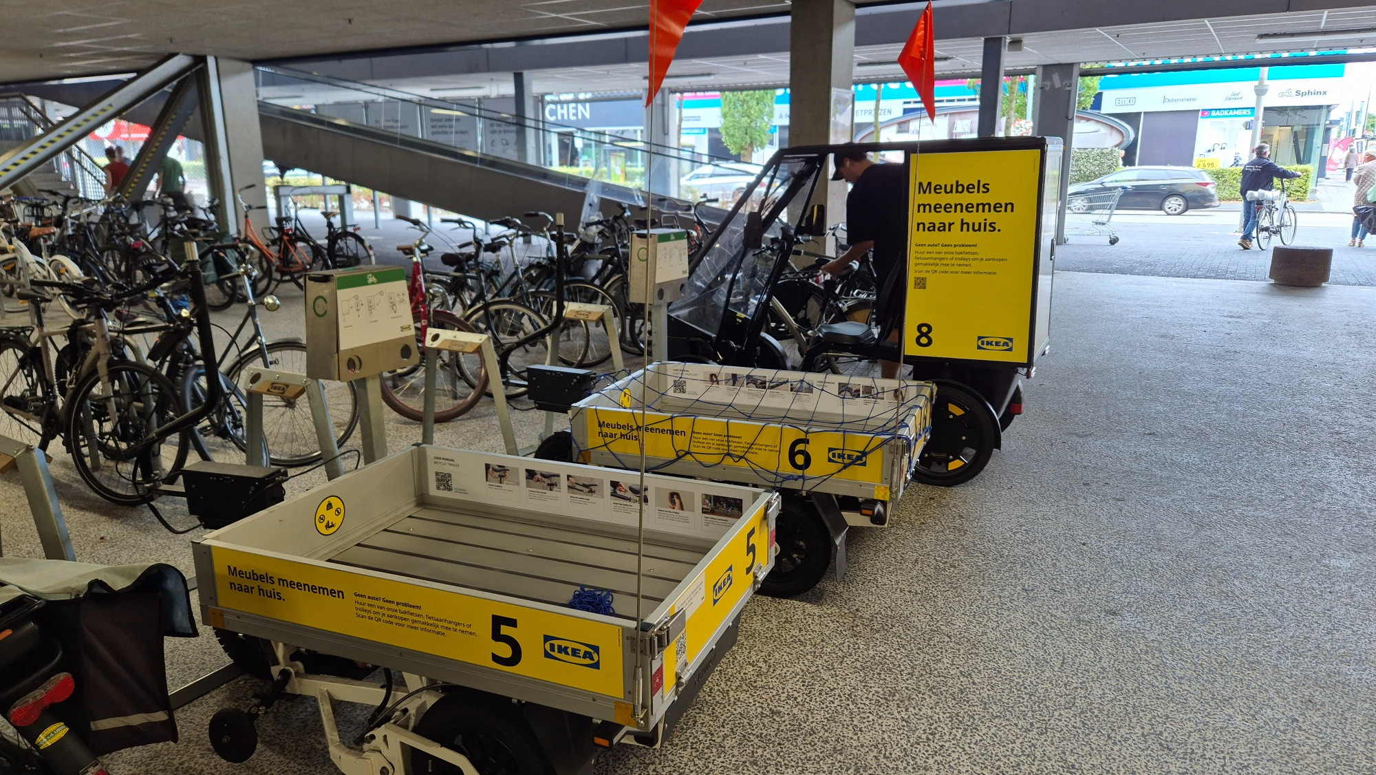 
Photo of two IKEA e-bike trailers and a covered IKEA cargo bike. The following text appears on the sides:

"Take furniture home.

No car? No worries!

Rent one of our cargo bikes, bike trailers, or trolleys to easily transport your purchases. Scan the QR code for more information."

The inside of the e-bike trailers features images explaining how to attach the trailer to your bike.