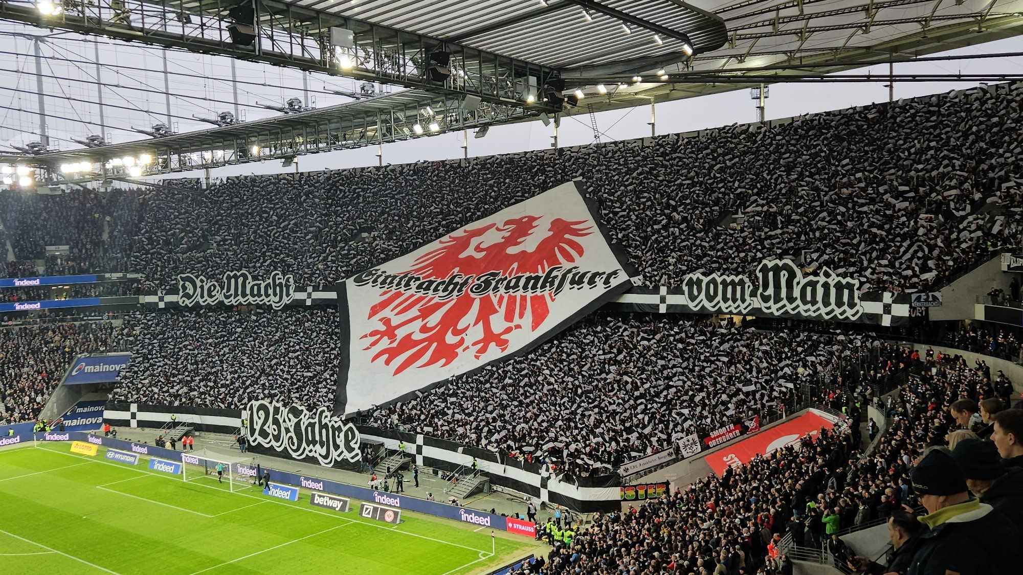 Nordwestkurve im Waldstadion. Die Fans mit schwarz/weissen Fähnchen. In der Mitte ein Karo mit dem alten Adler in Rot und dem Schriftzug Eintracht Frankfurt in Schwarz.
Dazu Banner mit der Aufschrift Die Macht vom Main 125 Jahre