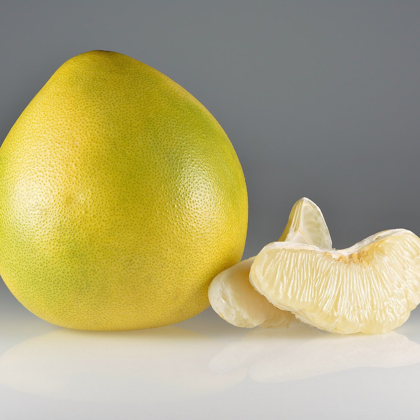 A imagem apresenta um pomelo inteiro e algumas de suas gomos separados. O pomelo inteiro é de formato oval, com casca amarela grossa e levemente rugosa. Ele está posicionado à esquerda da imagem, com a parte superior levemente inclinada para a direita. A casca apresenta um brilho sutil, indicando frescor.

À direita do pomelo inteiro, há três gomos separados. Os gomos são de cor branca e possuem uma textura suculenta e translúcida. Eles estão dispostos de forma a mostrar a divisão natural da fruta em segmentos. A polpa dos gomos é visível, com suas vesículas cheias de suco.

O fundo da imagem é cinza claro e uniforme, criando um contraste suave com as cores vibrantes da fruta. A iluminação é suave e difusa, destacando as formas e texturas do pomelo sem criar sombras muito marcadas. A imagem como um todo transmite uma sensação de frescor, naturalidade e apetitosidade.