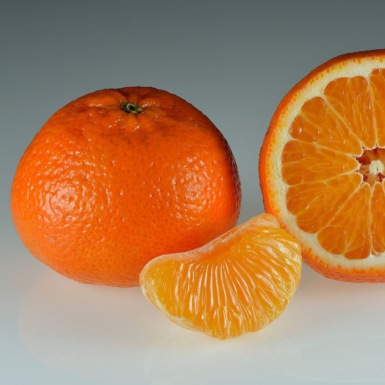 A imagem apresenta três tangerinas sobre um fundo cinza claro. A tangerina à esquerda está inteira, com sua casca alaranjada brilhante e textura ligeiramente rugosa. Seu formato é arredondado, com uma leve ponta na parte superior.
Ao lado da tangerina inteira, há uma tangerina cortada ao meio, revelando sua polpa suculenta e dividida em gomos. A polpa é de cor laranja vibrante e cada gomo contém pequenas vesículas cheias de suco. A casca da metade da tangerina também é visível, com sua parte interna branca e esponjosa contrastando com a parte externa alaranjada.
Completando a composição, há um gomo de tangerina separado, com sua forma curva e segmentos bem definidos. O gomo exibe a mesma cor vibrante e textura suculenta da polpa da tangerina cortada ao meio.