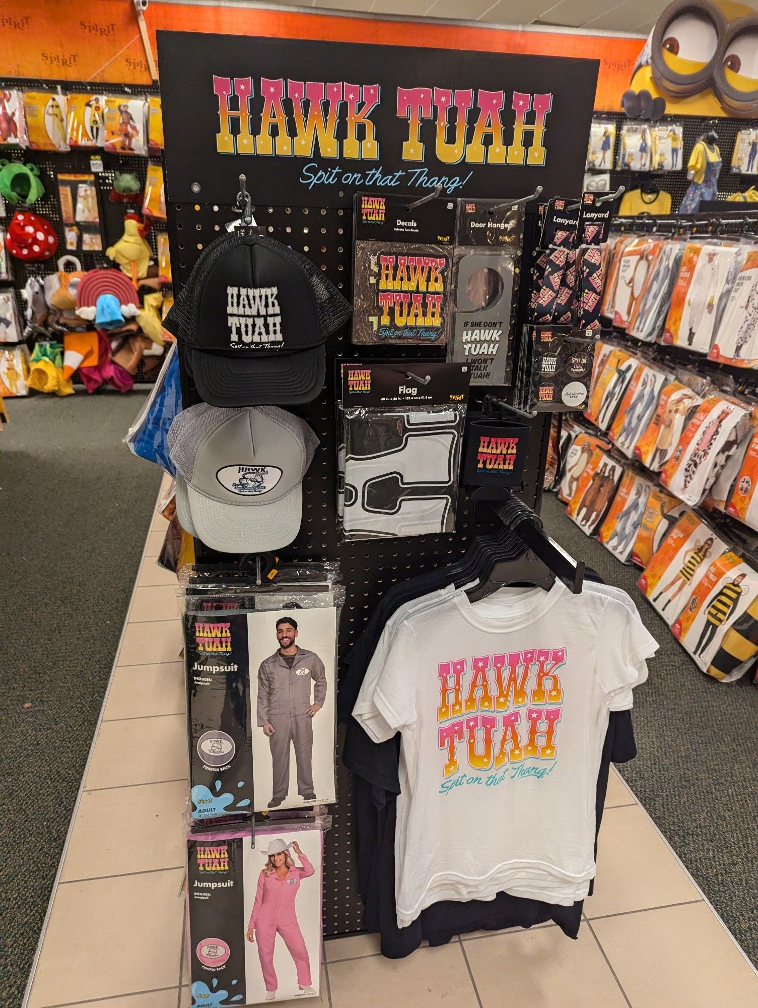 A display rack featuring merchandise based on the "Hawk Tuah" meme, including hats, T-shirts, jumpers, flags, buttons, lanyards, decals, and beer cookies. In the background are more traditional Halloween costumes.