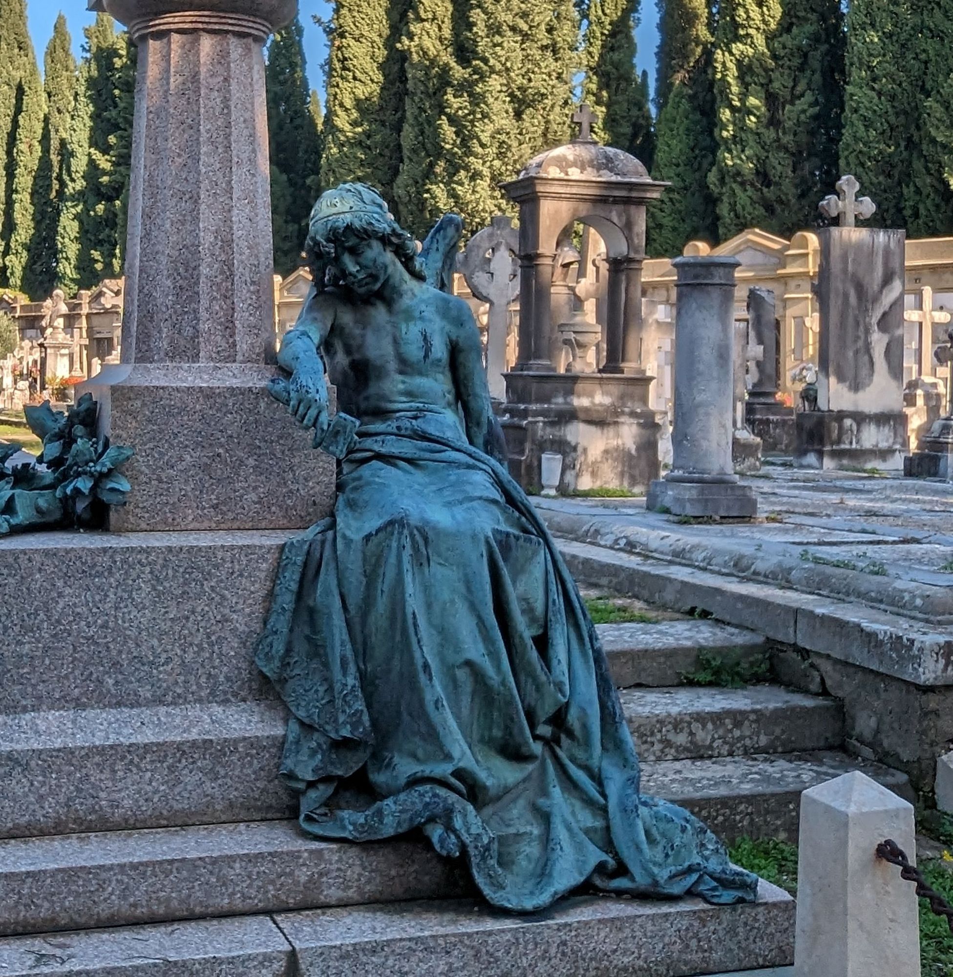 a bronze classical statue of a cherub in a long skirt holding a hammer, looking down in grief and exhaustion. the statue adorns a tomb at a graveyard in florence