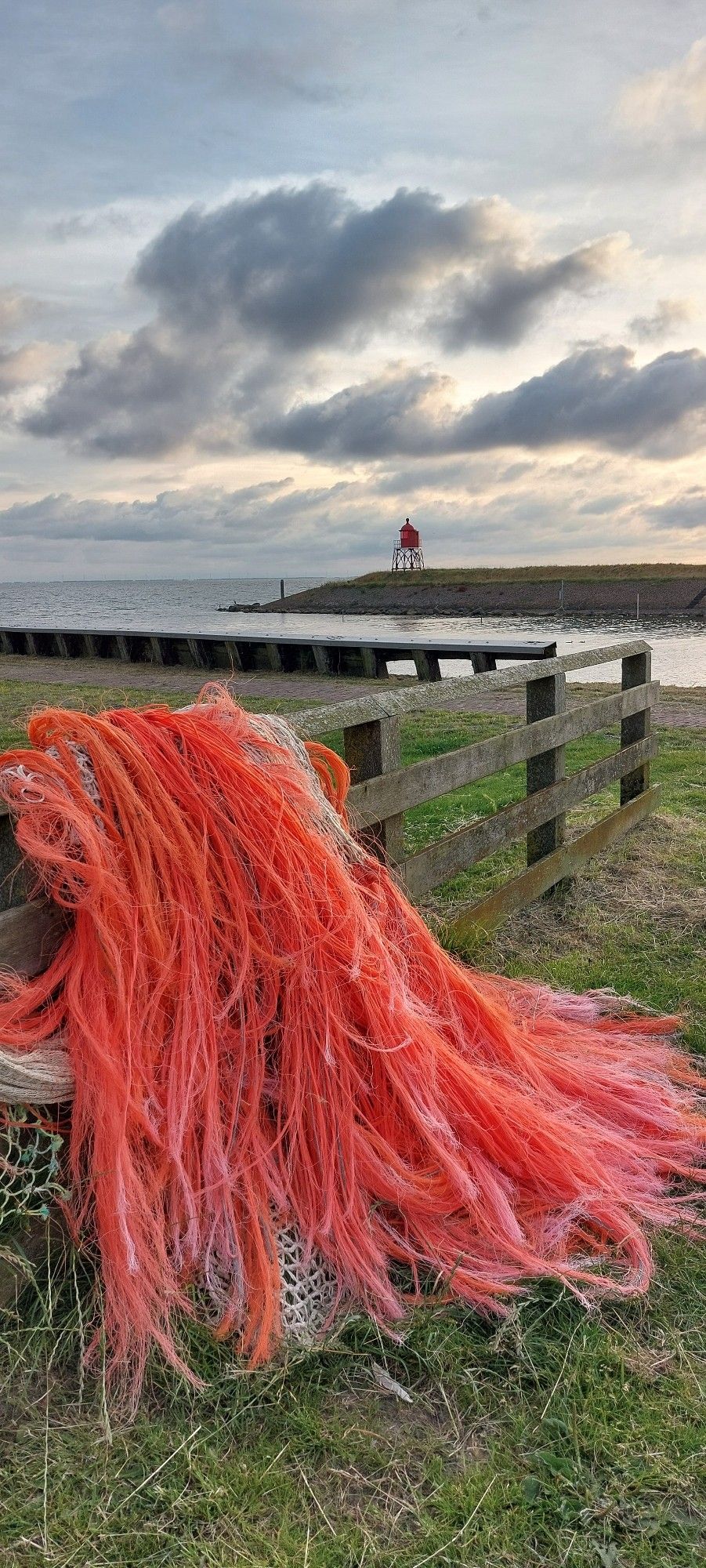 Een hek op het gras met een groot feloranje visnet erover. Verderop het water van de ingang van een haven met een klein rood baken erop. Daar weer achter het IJsselmeer en mooie wolken