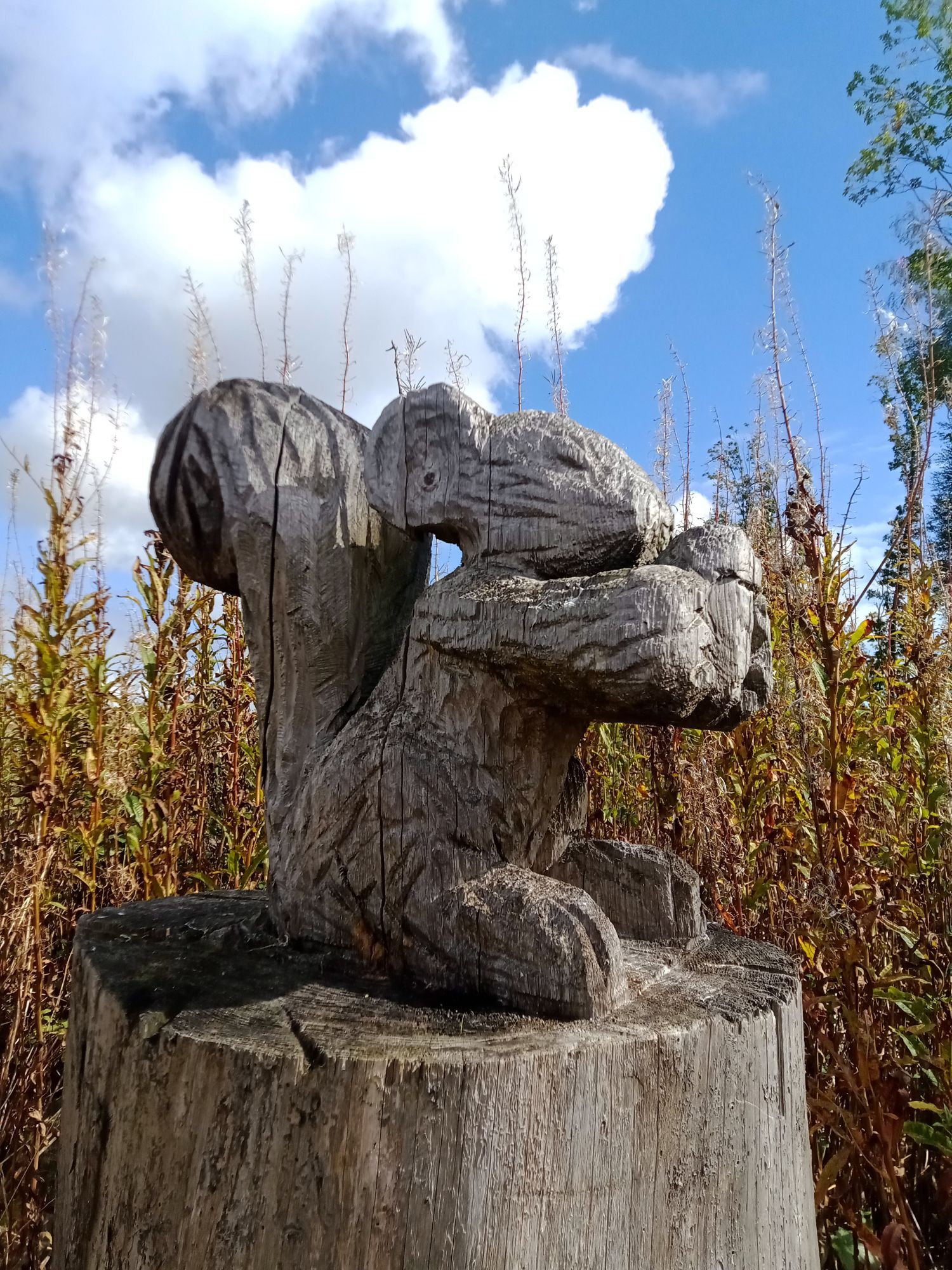 Eine von der Sonne angestrahlte und aus Holz geschnitzte Figur eines Eichhörnchens, das eine Eichel in ausgestreckten Pfoten hält. Dahinter herbstlich verfärbte Pflanzen und blauer Himmel mit Wolken.