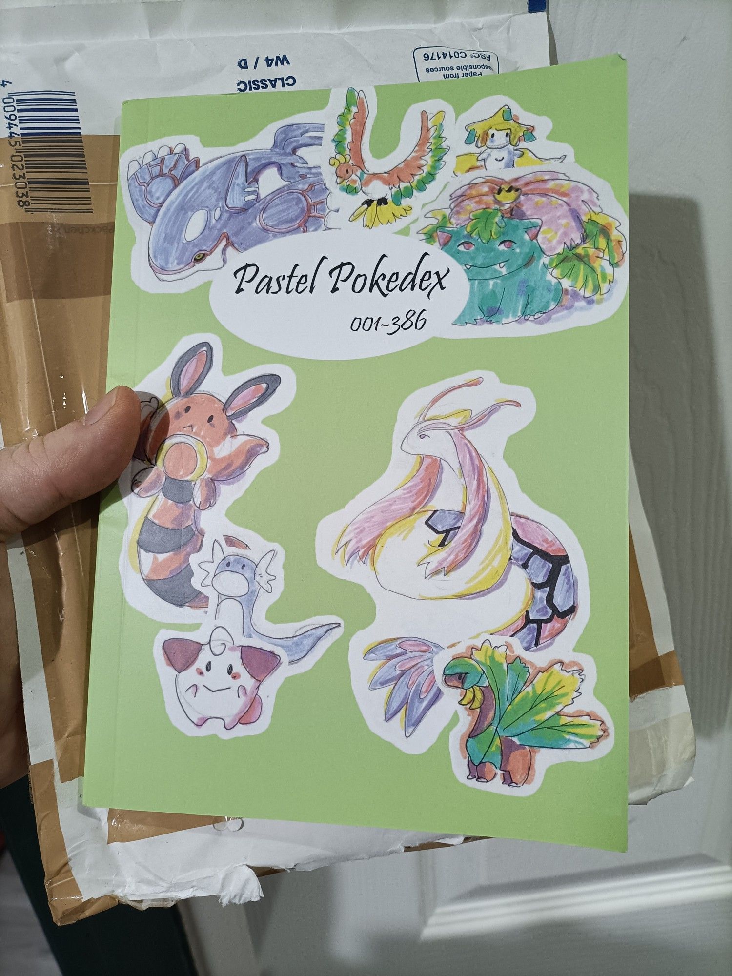 The front cover of a book called "Pastel Pokédex 001-386", covered in hand drawn Pokémon in a soft style. From top left going clockwise there are: Kyogre, Ho-oh, Jirachi, Venusaur, Milotic, Tropius, Cleffa, Dratini, and Sentret.