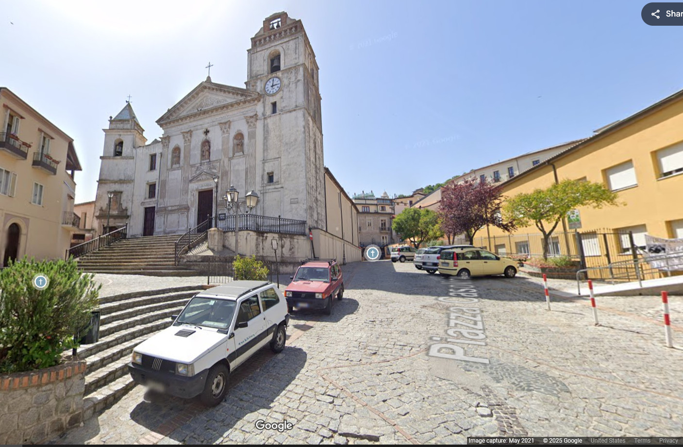 cobble stone piazza with a large church on the left. It has two bell towers. Some cars are parked in the piazza