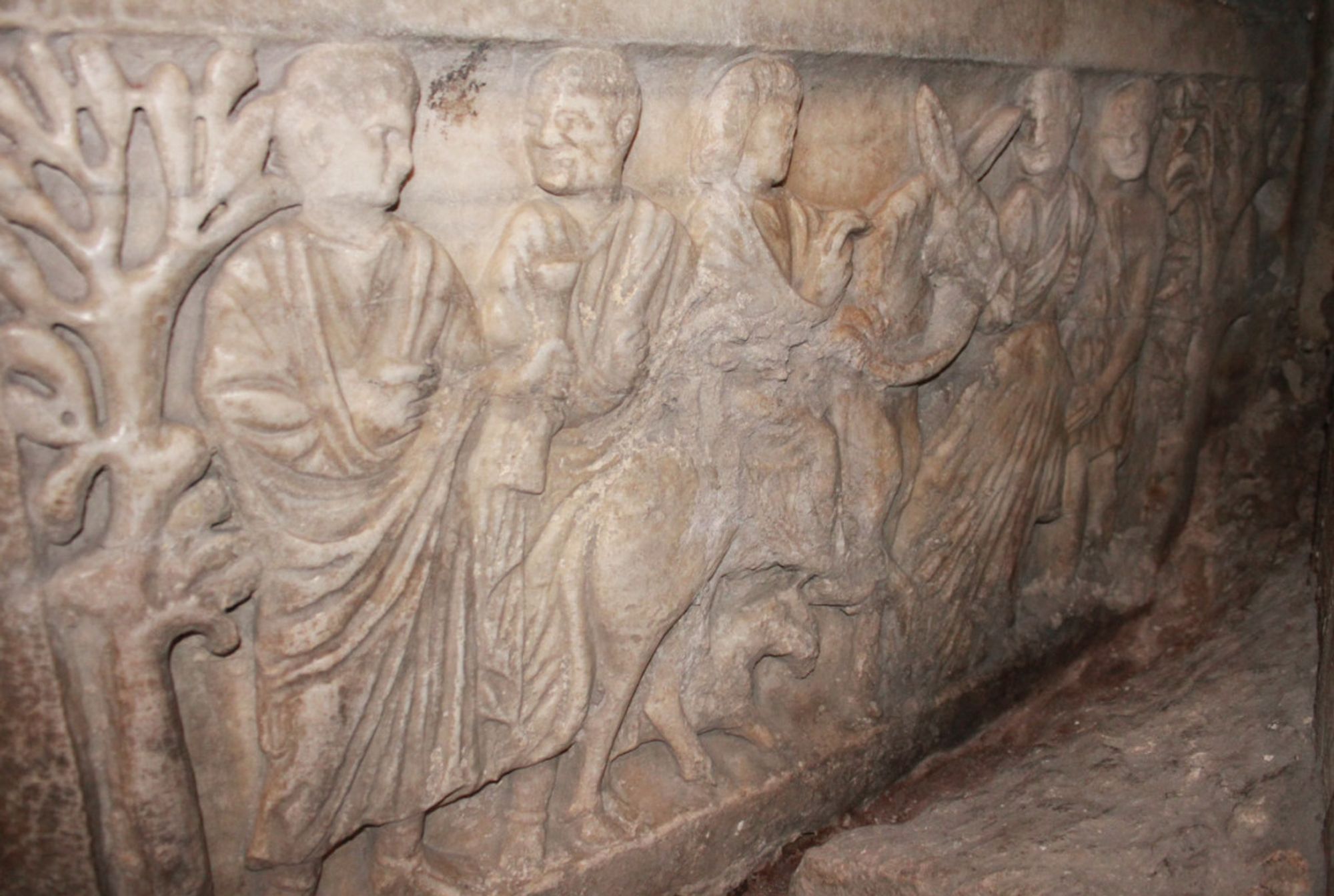 Marble relief showing Jesus riding on a donkey. Another man on the left stands under a tree talking to another man. In front of the donkey a man lays out a robe on the ground.