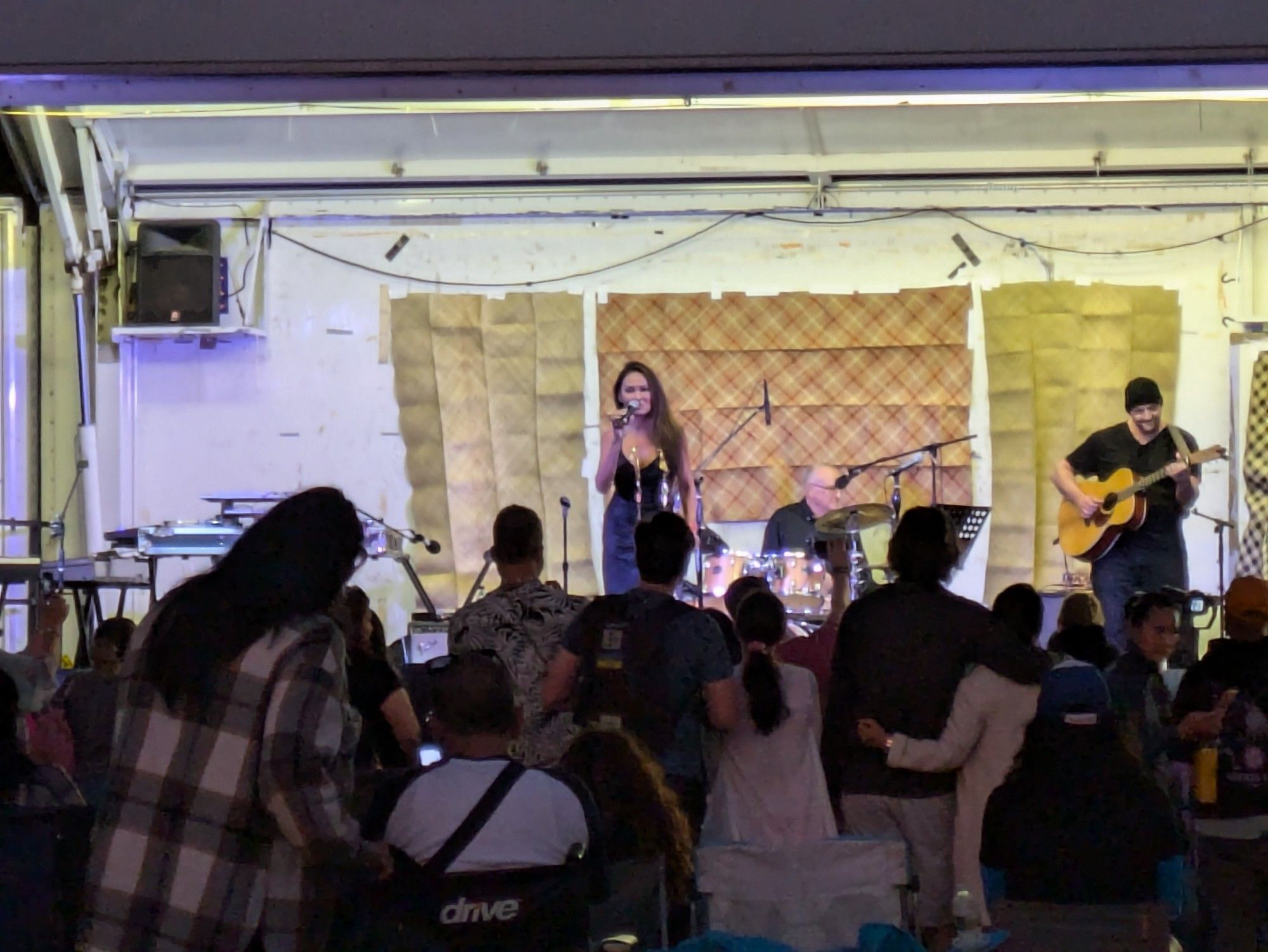 A photo of a ramshackle stage with a small audience standing in front. At center stage is Tia Carrere singing, while to her left someone is on guitar and behind them someone is on drums.