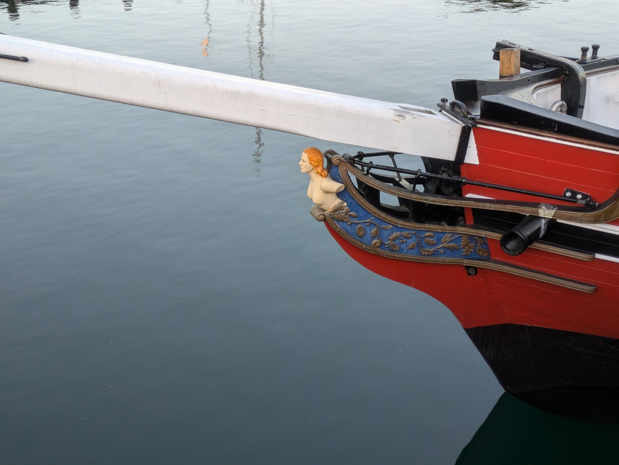 A photograph of the bow of a wooden ship. It has a bright red hull, an intricate blue and gold leaf pattern, and on the front a figurehead bust of a woman with light skin and orangish red hair. Yes, tits are out.