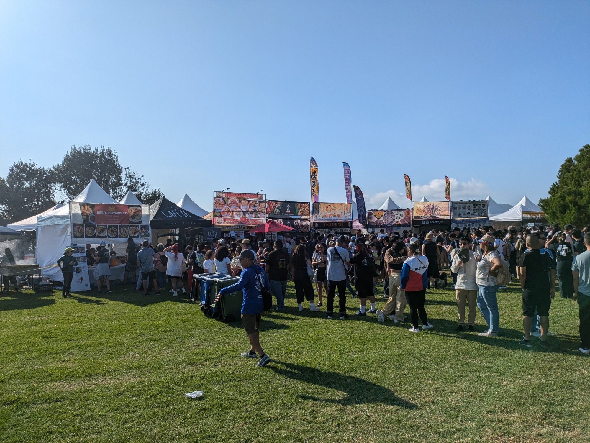 A photo of the food stalls at a festival. The sky is blue, the grass is green, and the lines are long. Very long.