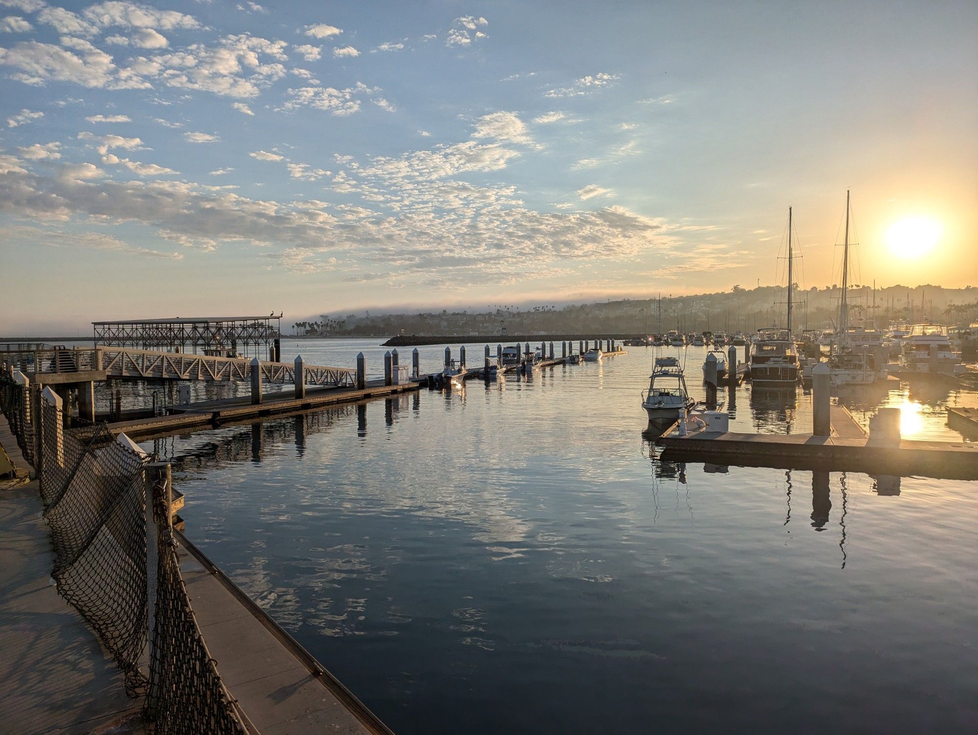 A landscape photograph taken on the edge of a marina. On the right you can see some boats at anchor, while a pier sticks out into the water in the left. In the distance the coast rises from the ocean, with the sun setting over the hill on the right and fog slowly devouring the land on the left. A scattering of clouds break up a light blue sky overhead.