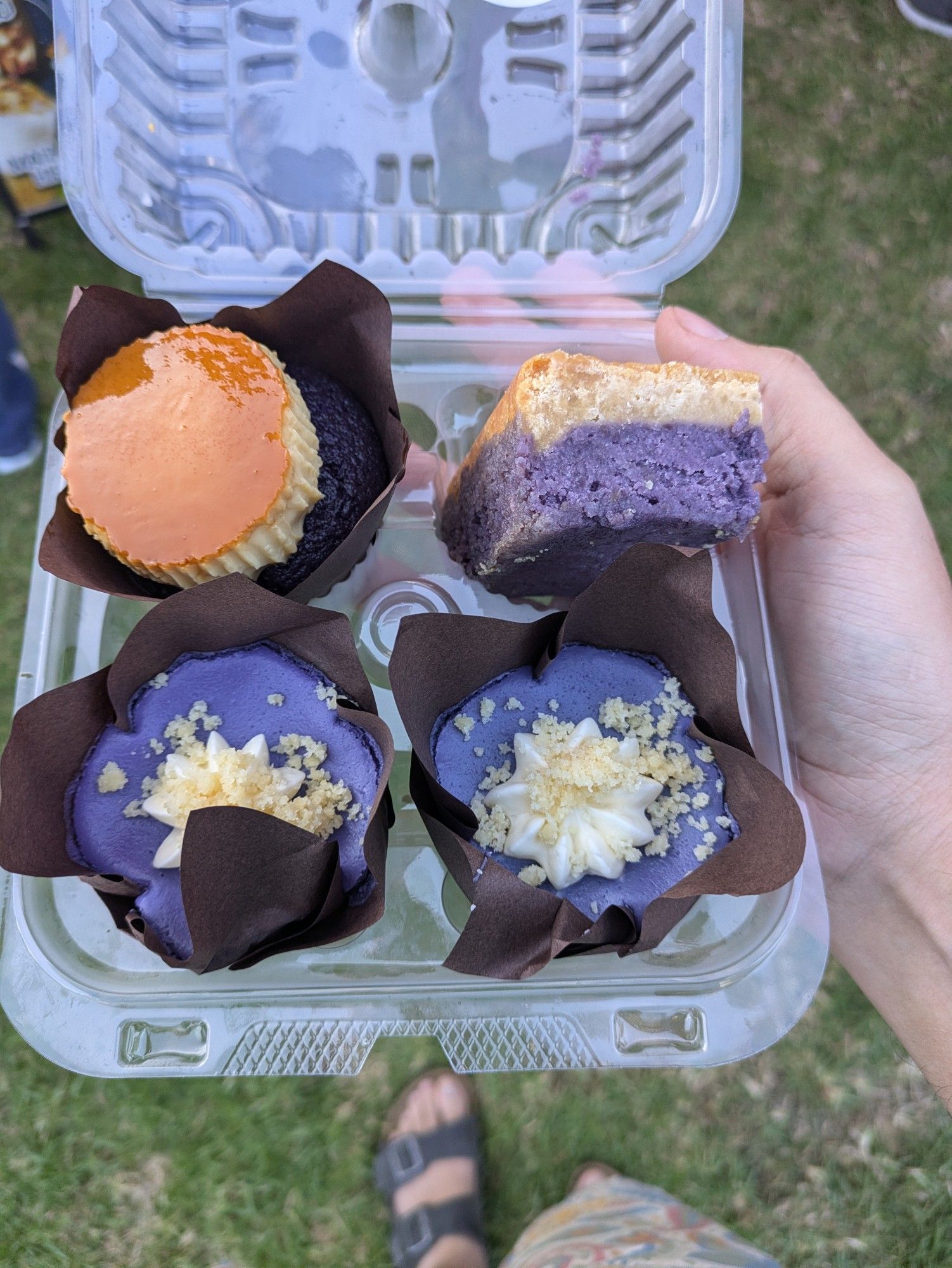 A plastic container containing four ube-flavored treats; one a cupcake covered in a small flan, one a ube butter bar, and two ube cheesecakes.