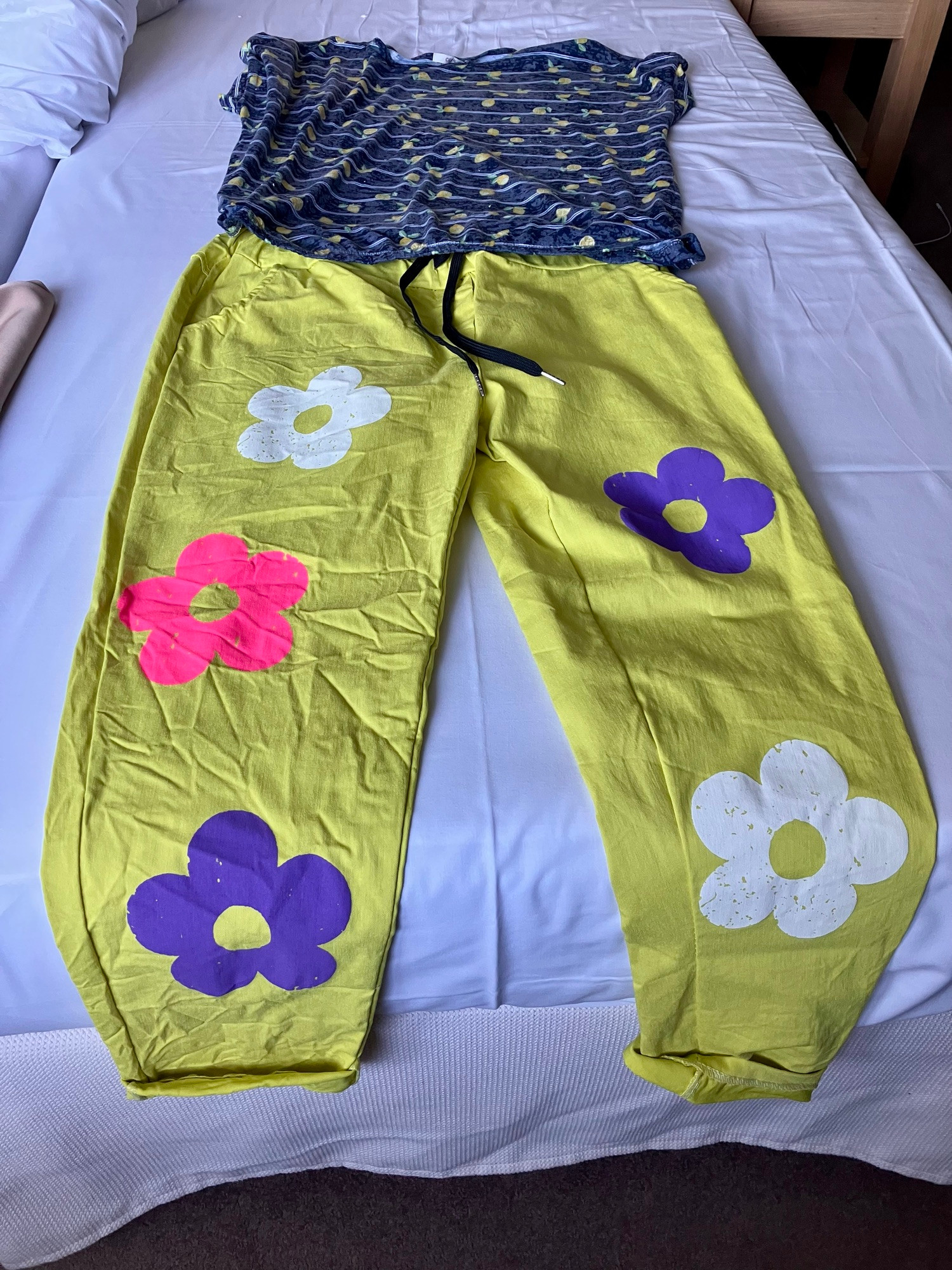 Ein lässiges Outfit, das auf einem Bett liegt, mit einem gestreiften blau-gelben Oberteil und einer leuchtend gelben Hose, mit pinken, weißen und blauen Blumen.
