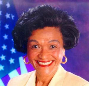 Ezola Foster, VP candidate on the 2000 Reform Party ticket. Photo of a late middle aged  Black Woman with a wide smile wearing gold earrings and a yellow suit jacket on a purple background with an American flag on the left side. https://en.wikipedia.org/wiki/Ezola_Foster