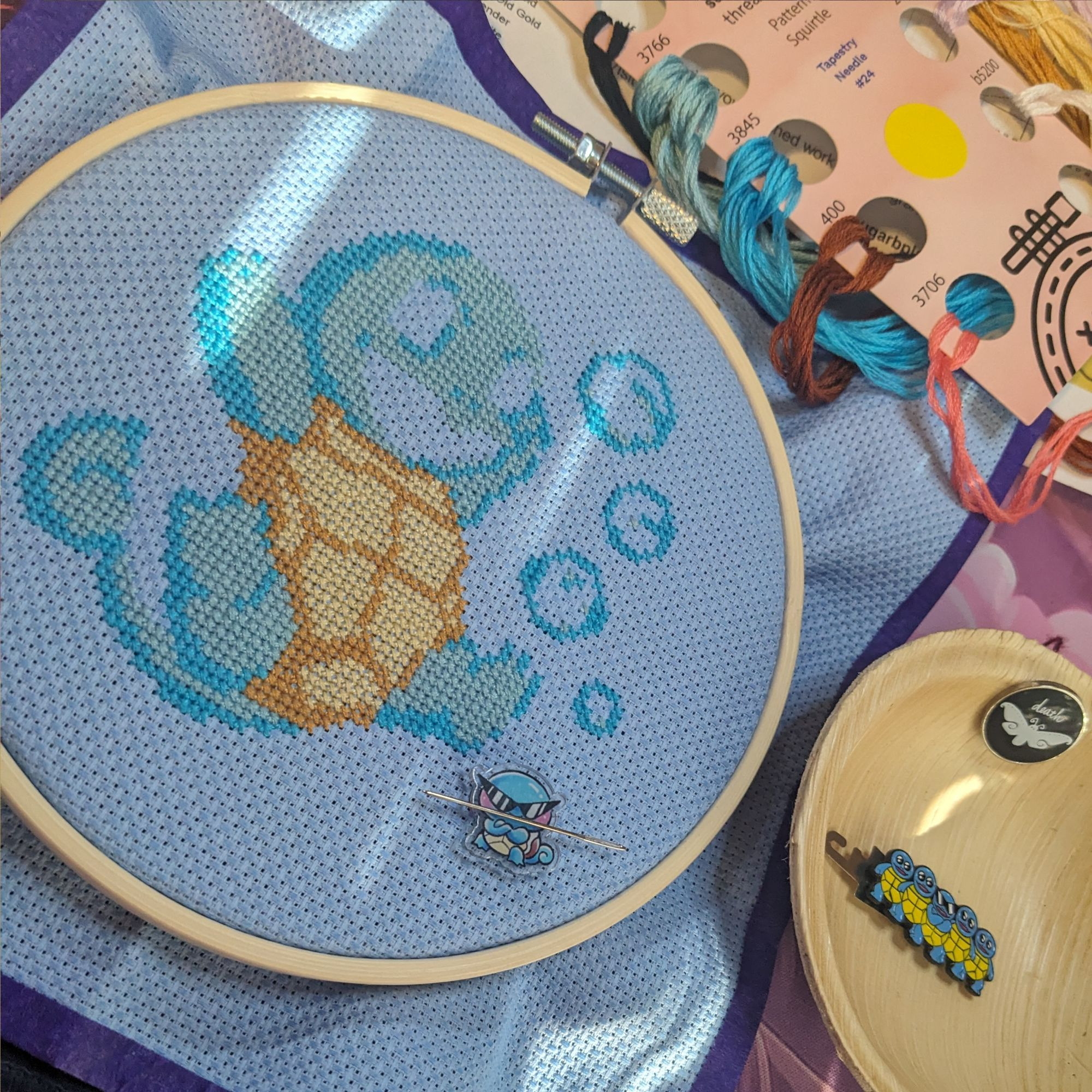 An unfinished Squirtle cross stitch project with a Squirtle needle minder and the Squirtle gang as the my threader.