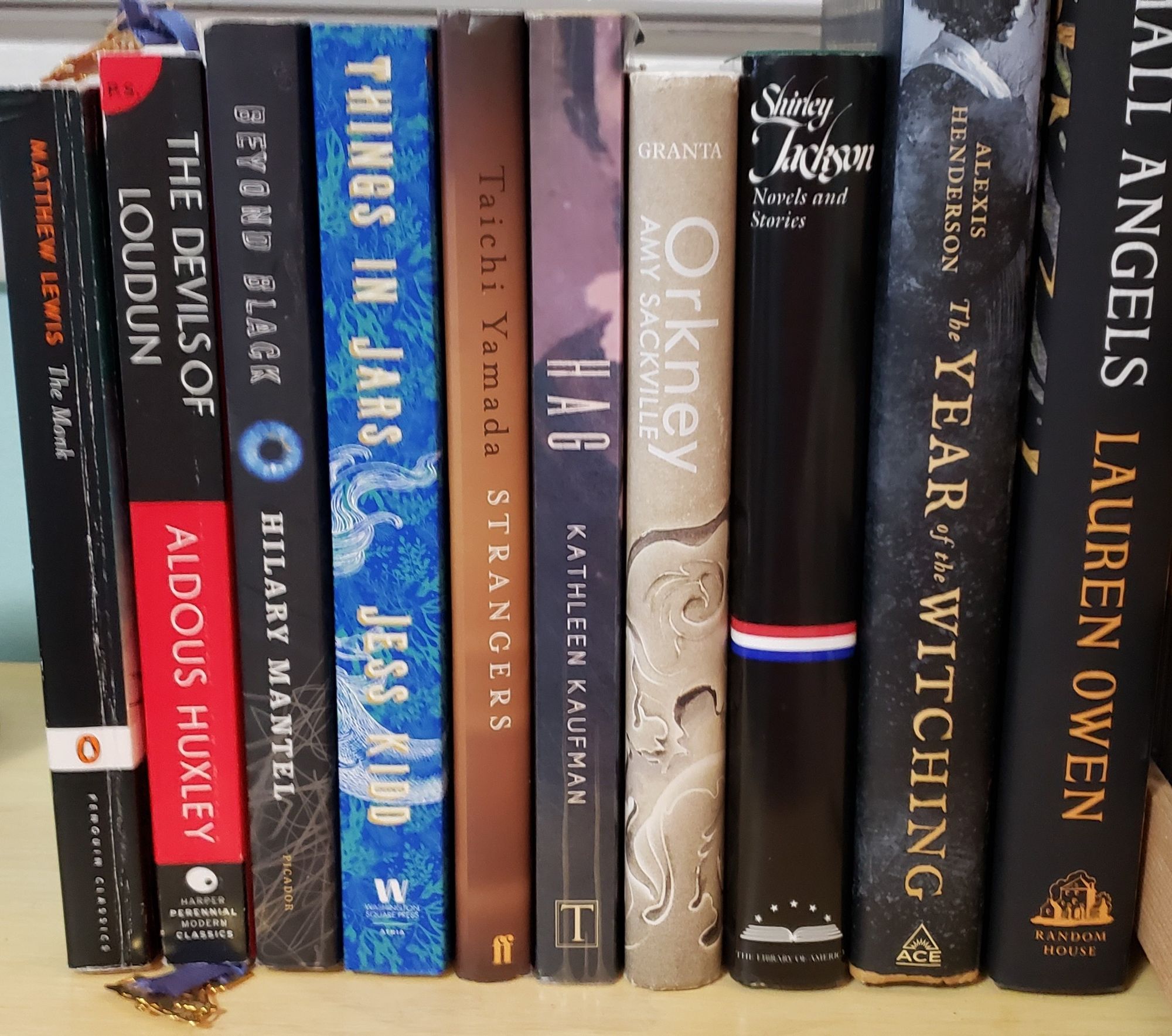 A stack of books on a shelf. Left to right; The Monk, Matthew Lewis; The Devils of Loudun, Aldous Huxley; Beyond Black, Hilary Mantel; Things in Jars, Jess Kidd; Strangers, Taiichi Yamada; Hag, Kathleen Kaufman; Orkney, Amy Sackville; a Library of America edition of Shirley Jackson novels; The Year of the Witch, Alexis Henderson; and Small Angels, Lauren Owen