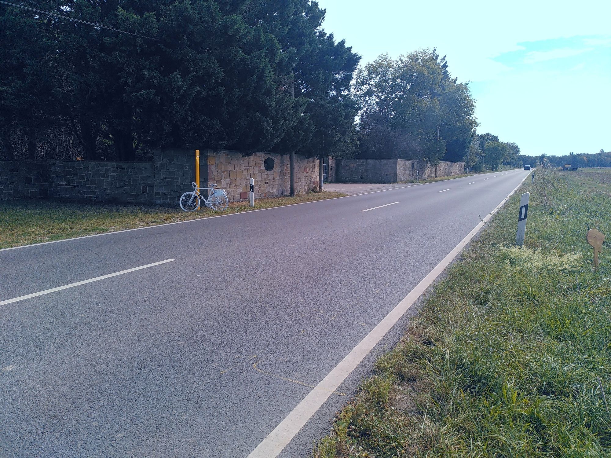 Straße mit Ackerfläche rechts und Bebauung links auf der linken Straßenseite steht ein lackiertes GHOSTBIKE an einen Poller angeschlossen.