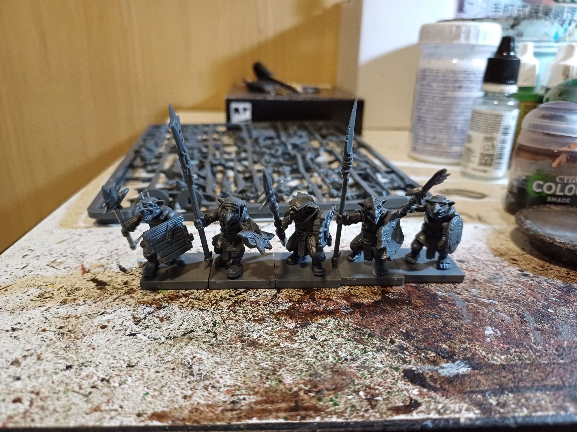 Photo: 5 goblin spearmen on my painting table by mantic games, behind them are two sprues of additional parts for five more warriors; various paint and glue bottles are also visible