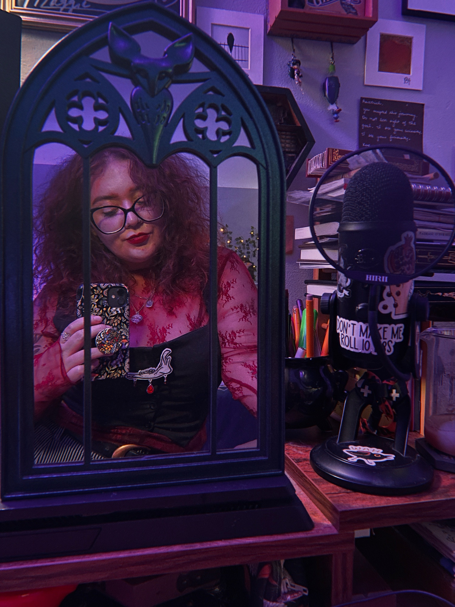 A mirror selfie of Lis in a mirror that looks like a stained glass window, on a desk with recording equipment. Wearing a red lace top, black vest, looking down.