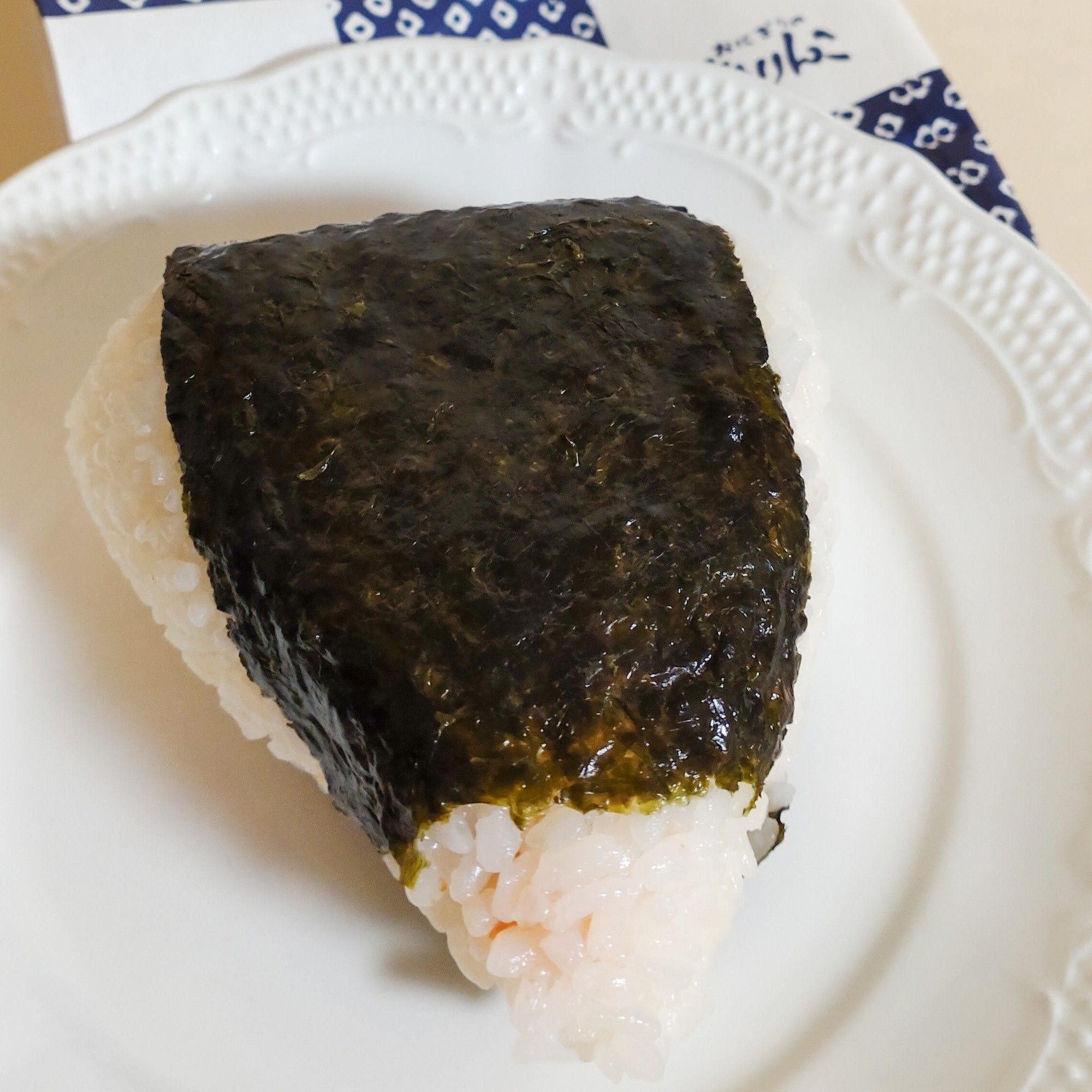 海苔の巻かれた三角おにぎりが白い皿の上に乗っている。ご飯の部分はサーモンのオレンジ色が透けて見える。皿の奥にはおにぎりのありんこの紙袋が僅かに見えている。