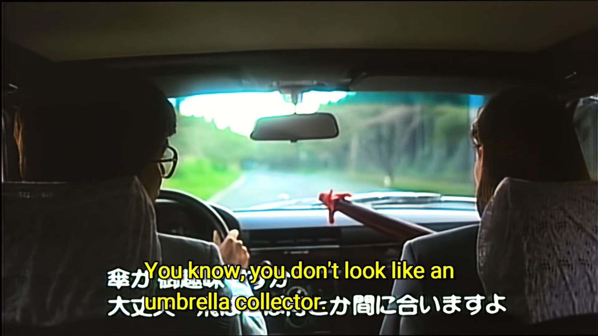 Frame from 【丹波哲郎の大霊界 死んだらどうなる】(Tetsuro Tamba's Great Spirit World: What Happens When You Die). A man driving a car is telling his woman passenger, who's holding an umbrella, "You know, you don't look like an umbrella collector."