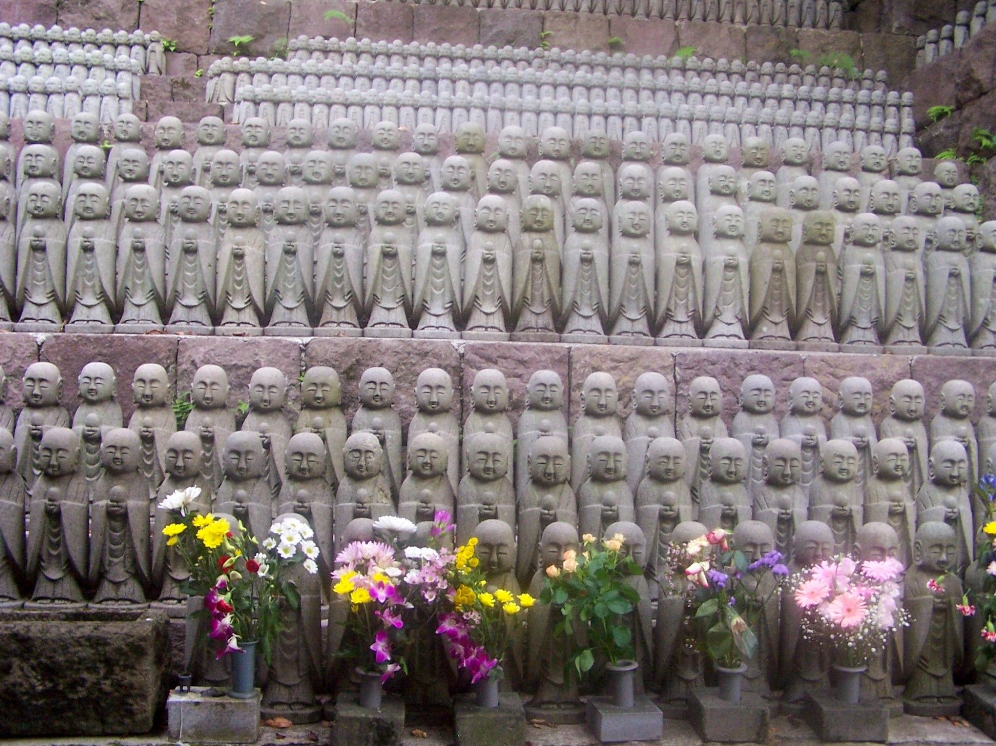 Kleine Steinfiguren in einheitlicher Größe und Ausführung stehen eng aneinander in endlosen Reihen.
Davor eine viele frische Blumensträuße.