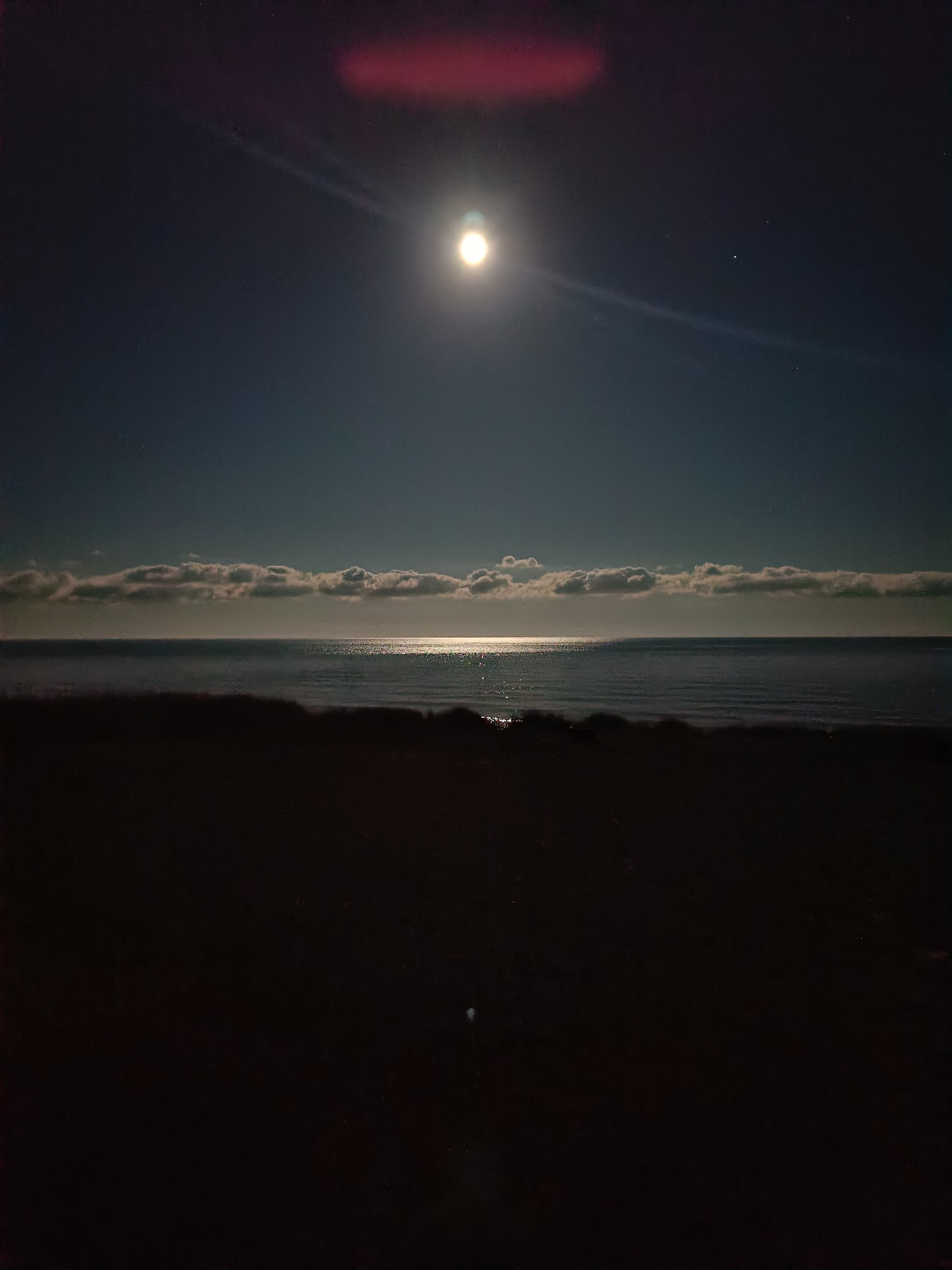 An almost full moon over the sea. There's a bright reflection of the moon on the water, and a gradient of the sky from pale grey-blue at the horizon to a black above the moon. There's a line of dramatically lit clouds a bit over the horizon, and over the moon there's a wide fuzzy red horizontal line. Possibly an artifact introduced by a camera struggling with too strong contrasts. Possibly an UFO.