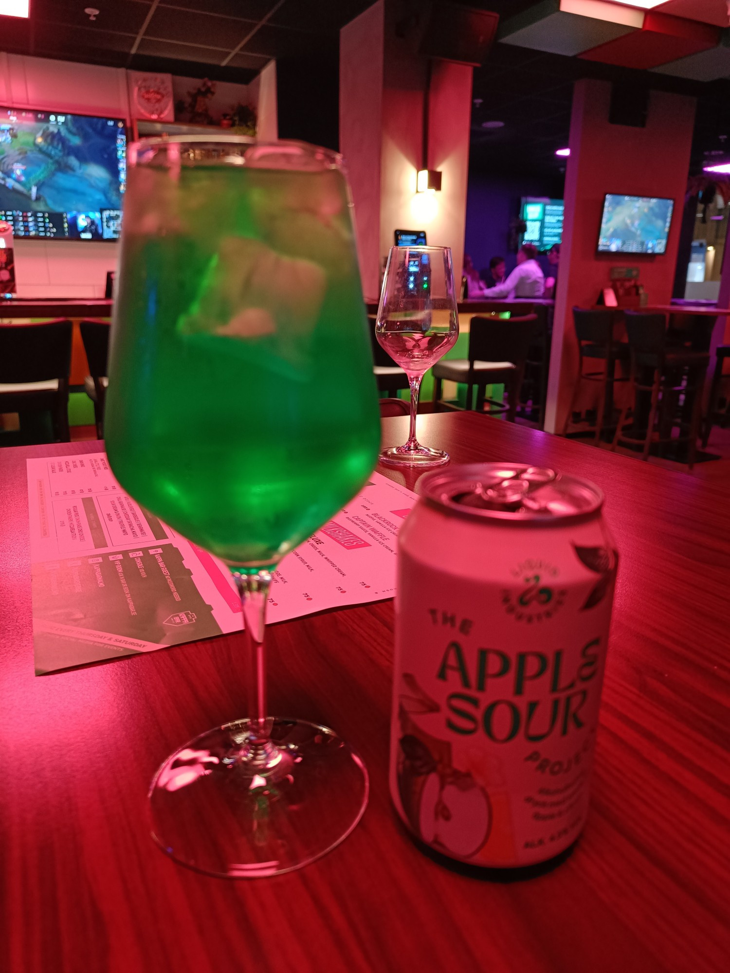 The interior of a bar with screens in the walls showing various games, bathed in peach and lilac lighting. 
In the foreground, an elegant stemmed glass with a vividly green liquid in.