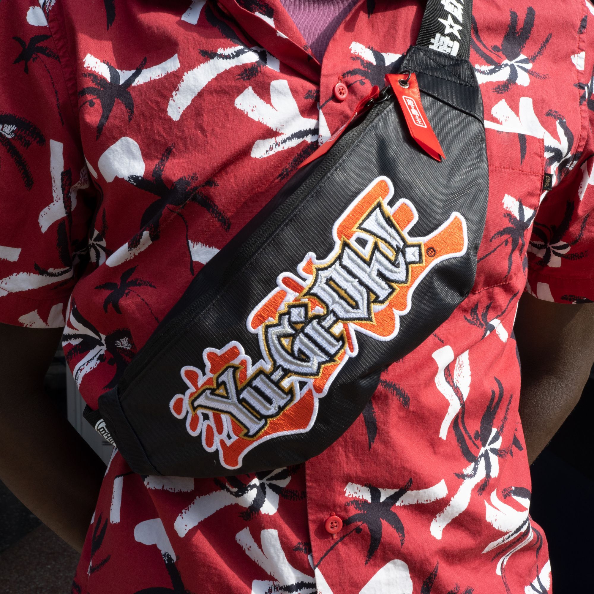 A photo of someone wearing the Yu-Gi-Oh! Fanny Pack across their chest. It is all black with printed elements on the strap. There is a Red Ribbon zipper pull and a large Yu-Gi-Oh! patch in the printed on the front.