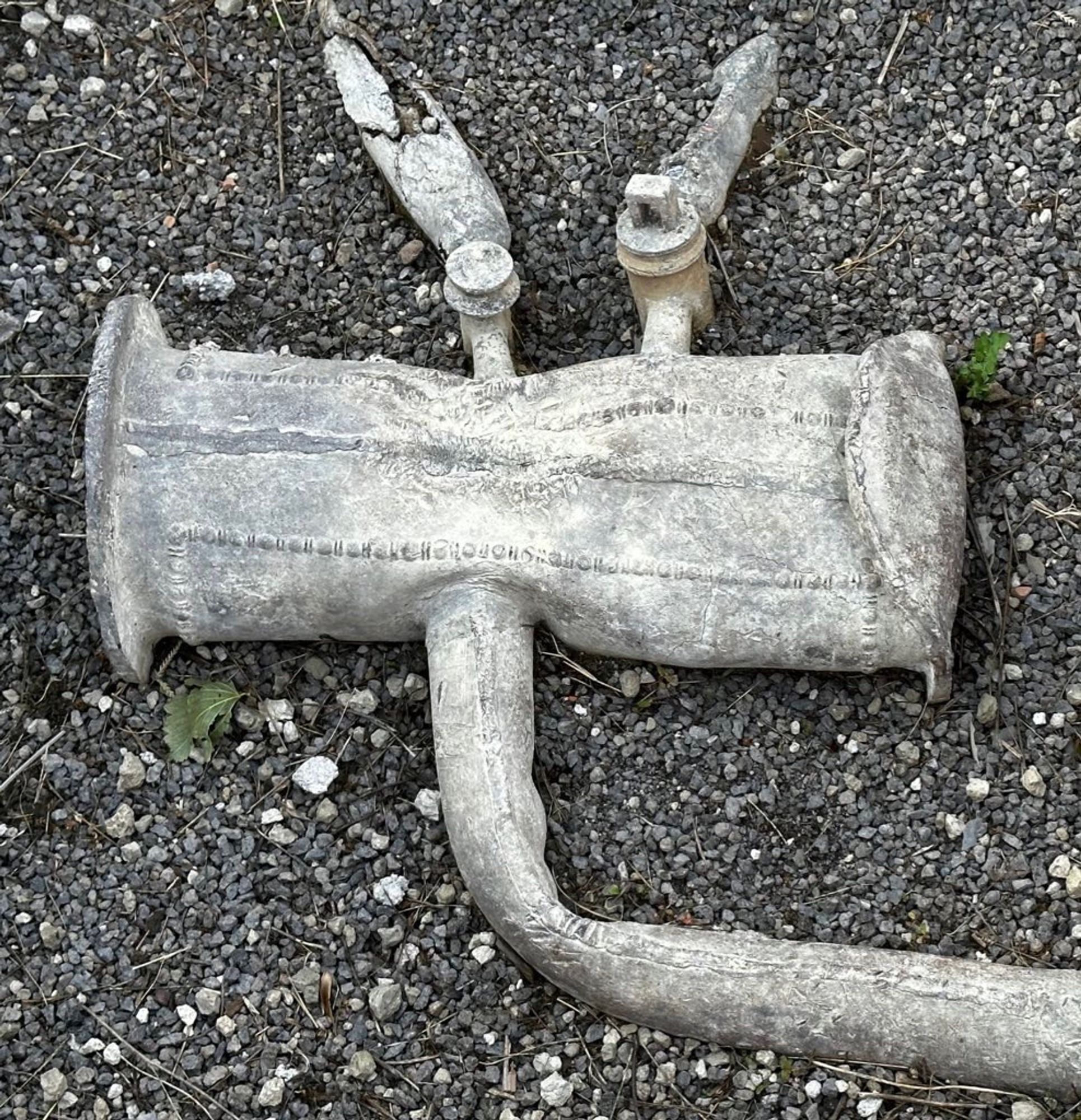 A lead reservoir in the shape of a drum with beaded decoration running along its length. It’s slightly squashed thanks to the eruption. From the right side of the image a single lead pipe enters the reservoir and two thinner pipes exit the drum on the far side. Both these smaller pipes have valves on them near the point of contact with the reservoir.