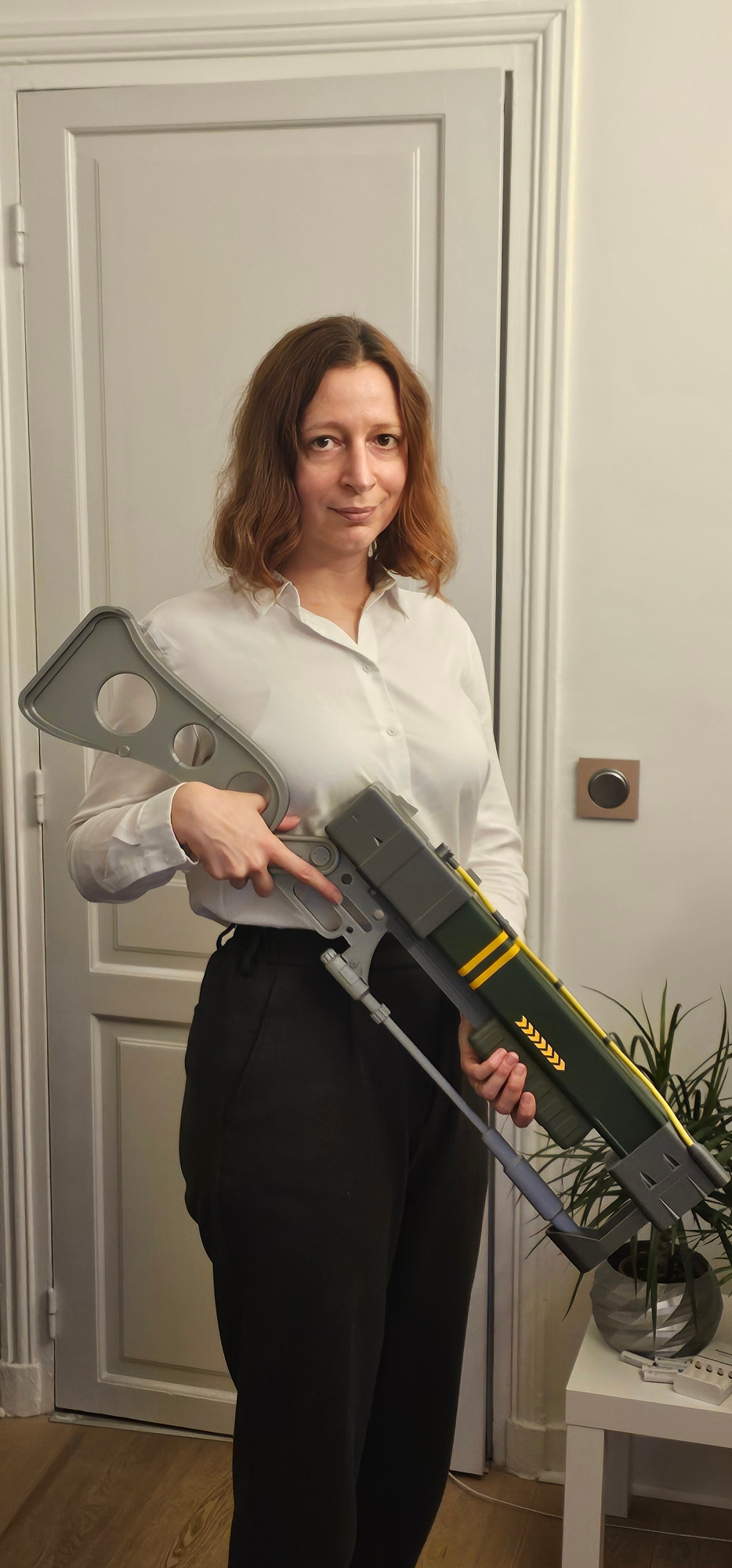 Me standing in my living room dressed in work clothes with a 3D printed recreation of a Fallout laser rifle. A cowering plant can be seen in the background.