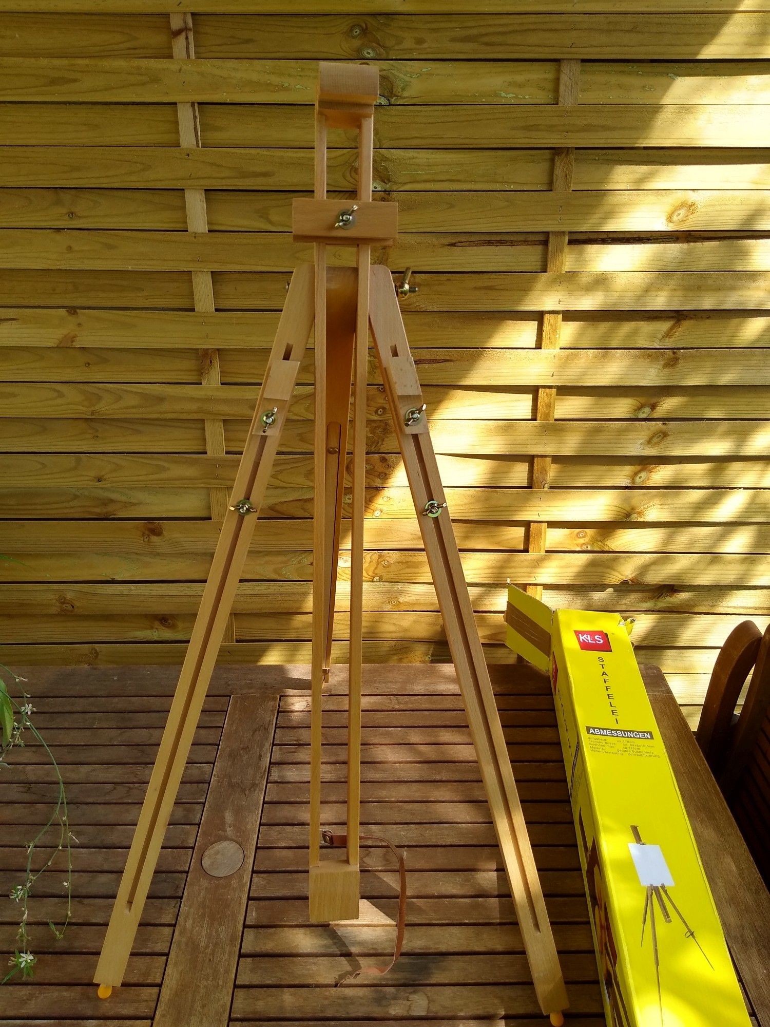 Staffelei aus Massivholz steht auf einem Holztisch, im Hintergrund eine Palisade.
Neben der Staffelei liegt die gelbe Verpackung.