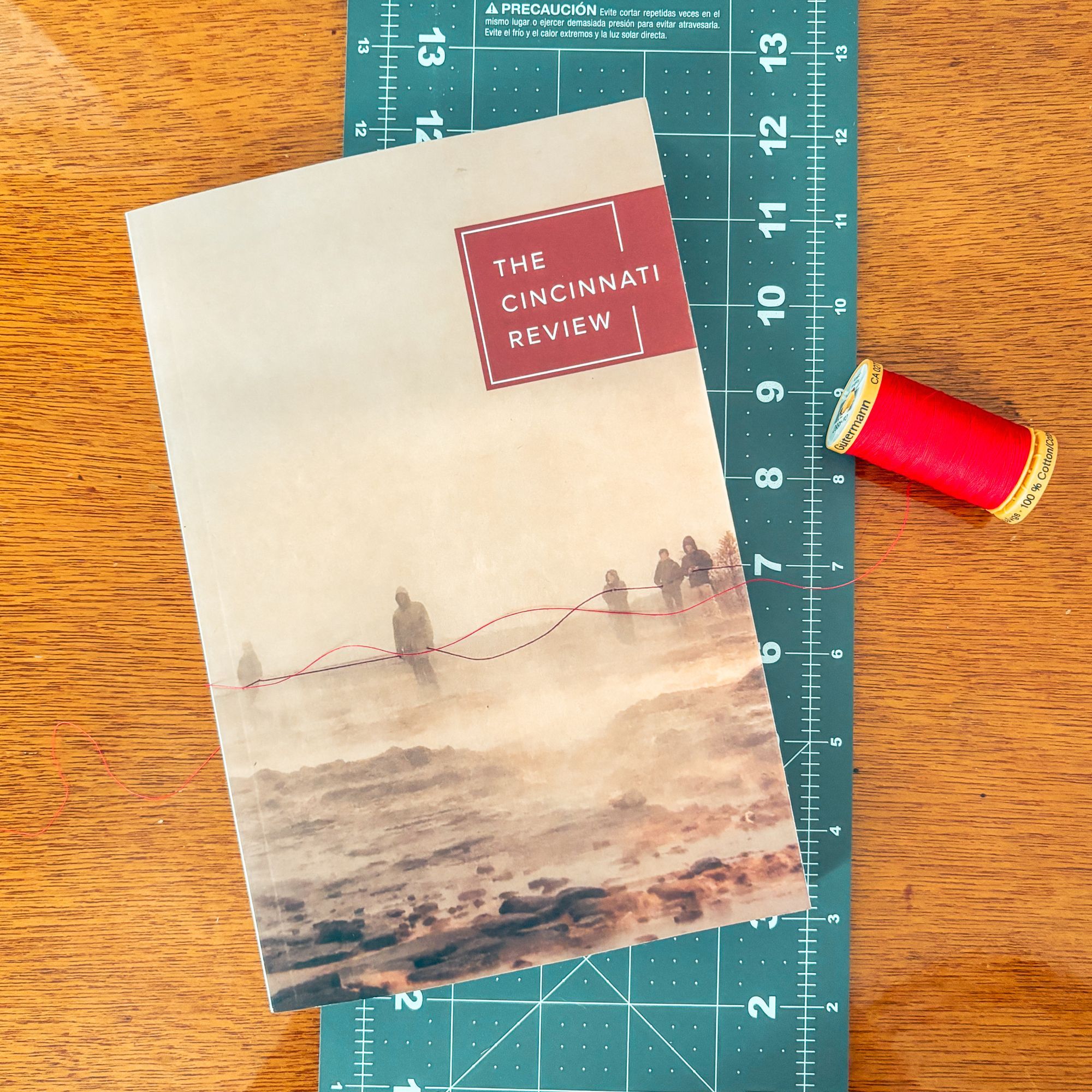 Issue 21.1 lying on a cutting mat (for sewing) next to a spool of red thread that is draped across the cover to match the image, which features figures all holding onto a thin red thread in an apocalyptic landscape.