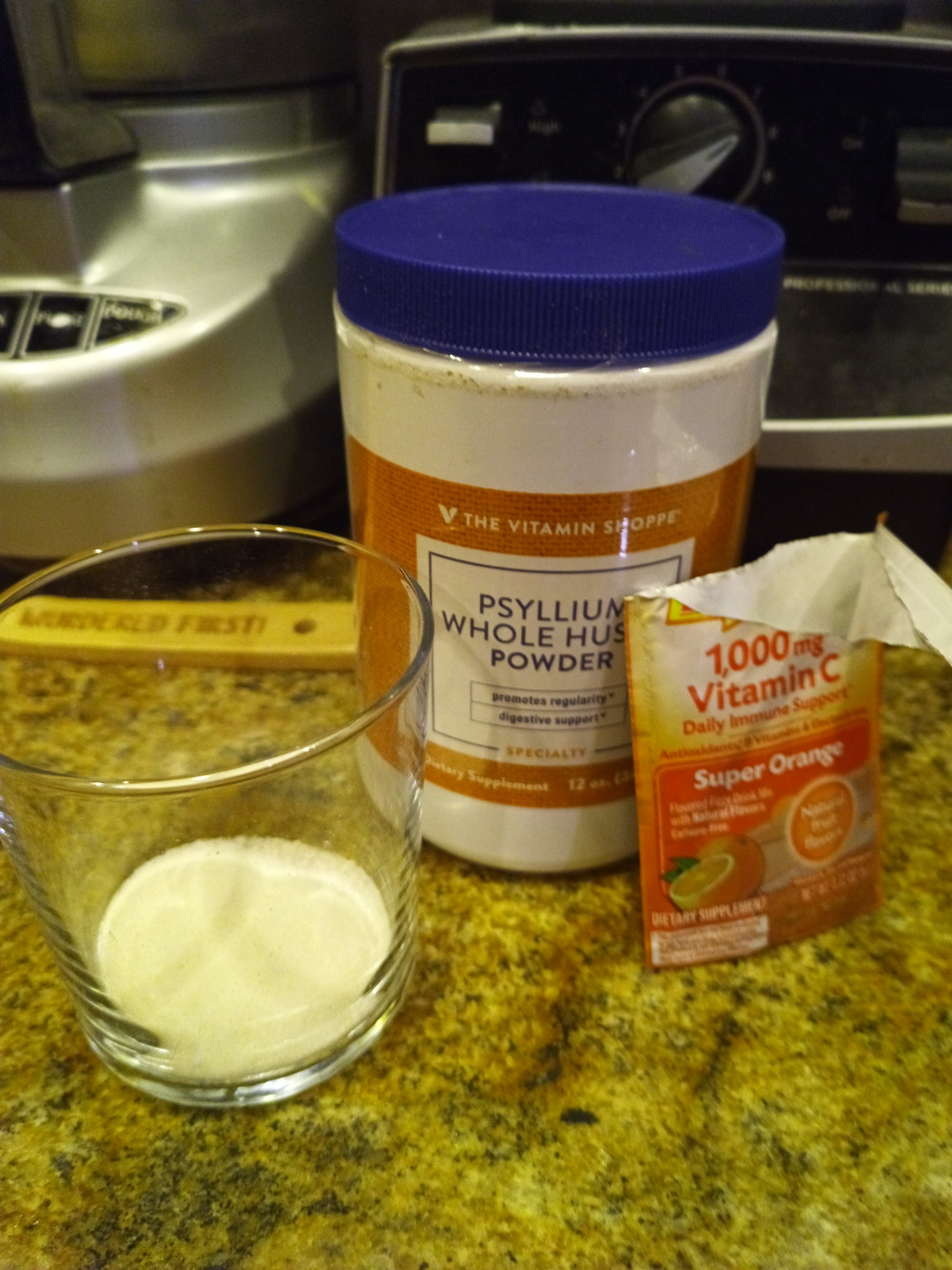Photo of a glass with two different types of powders mixed together in the bottom, next to a tub of psyllium powder and a ripped-open packet of emergen-c Super Orange, because photographer is a middle-aged person with a cold