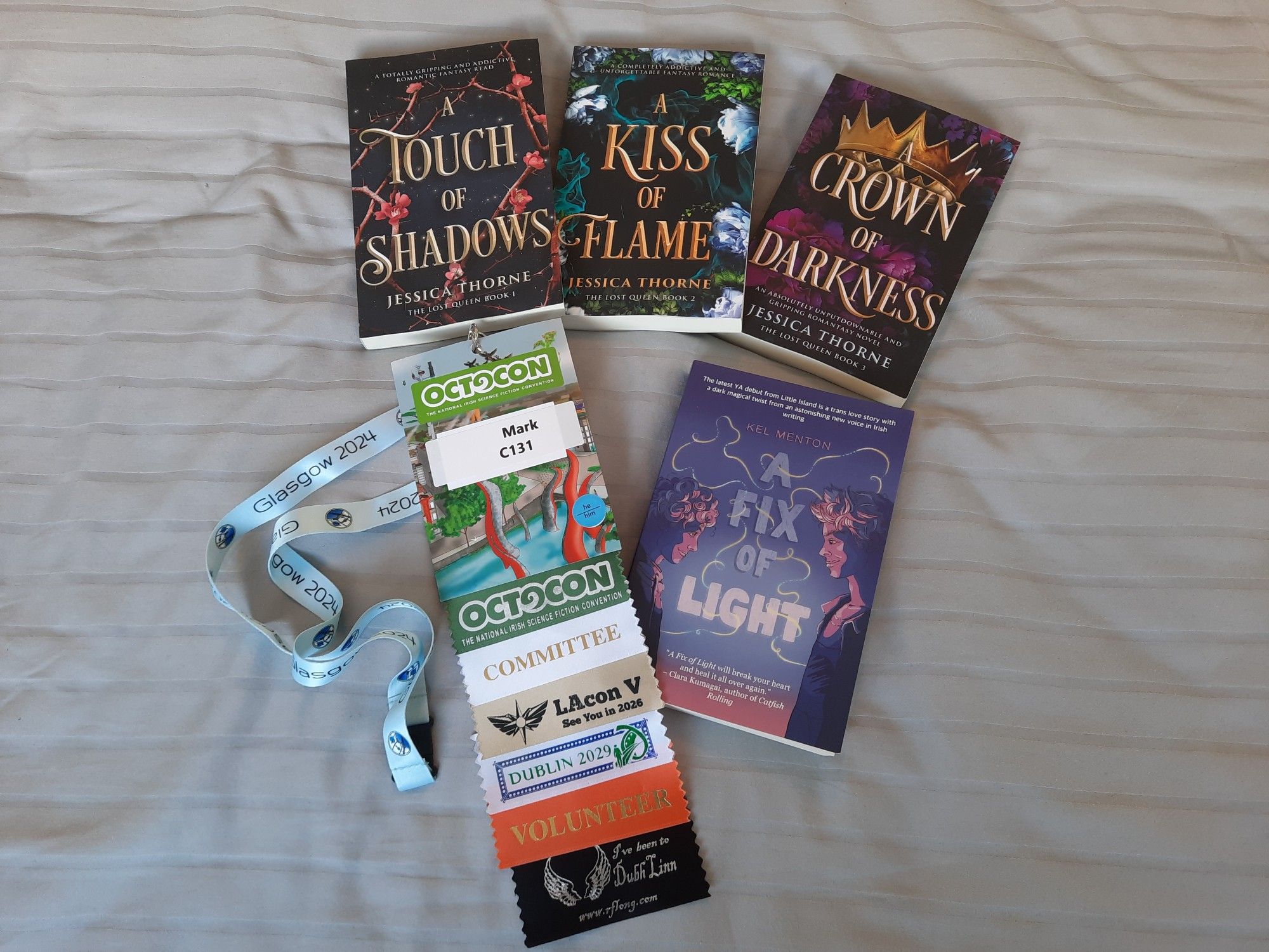 On a bed with grey sheets is my Octocon badge with Glasgow 2024 lanyard. Along with this is Jessica Thorne's The Lost Queen trilogy, A Touch of Shadows, A Kiss of Flame and A Crown of Darkness as well as Kel Menton's ARC copy of A Fix of Light.