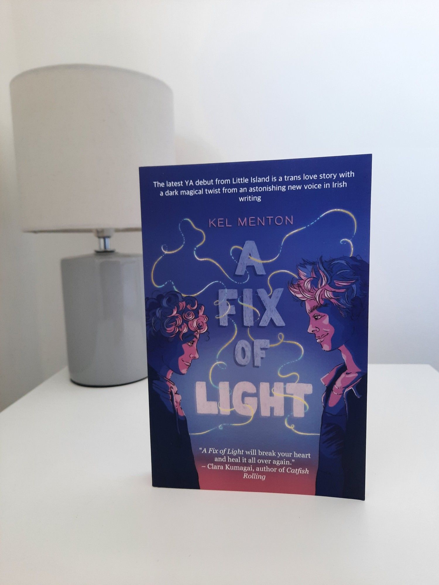 On a white bedside locker is Kel Menton's ARC copy of A Fix of Light in front of a grey lamp.