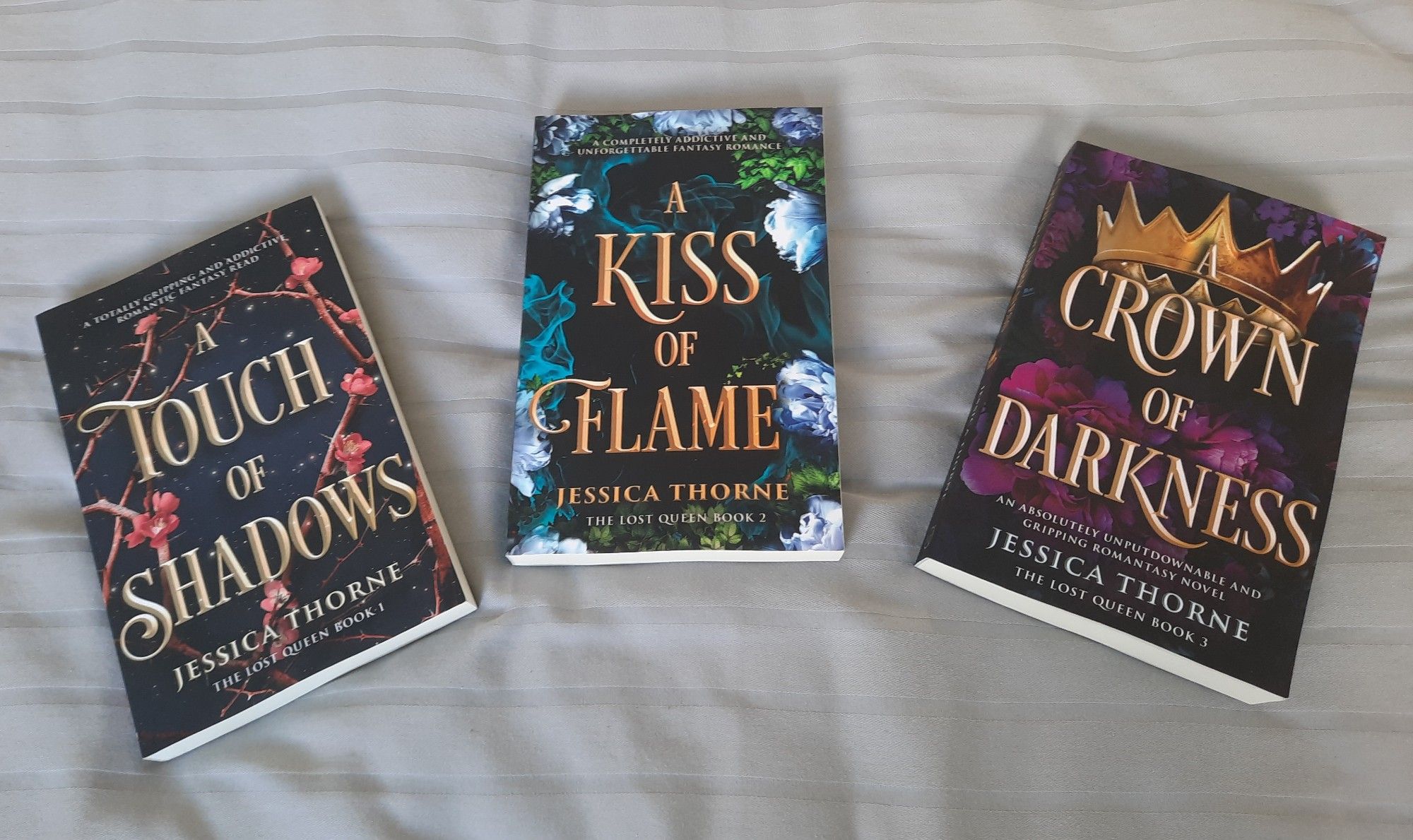 On a bed with grey sheets is Jessica Thorne's The Lost Queen trilogy, A Touch of Shadows, A Kiss of Flame and A Crown of Darkness.