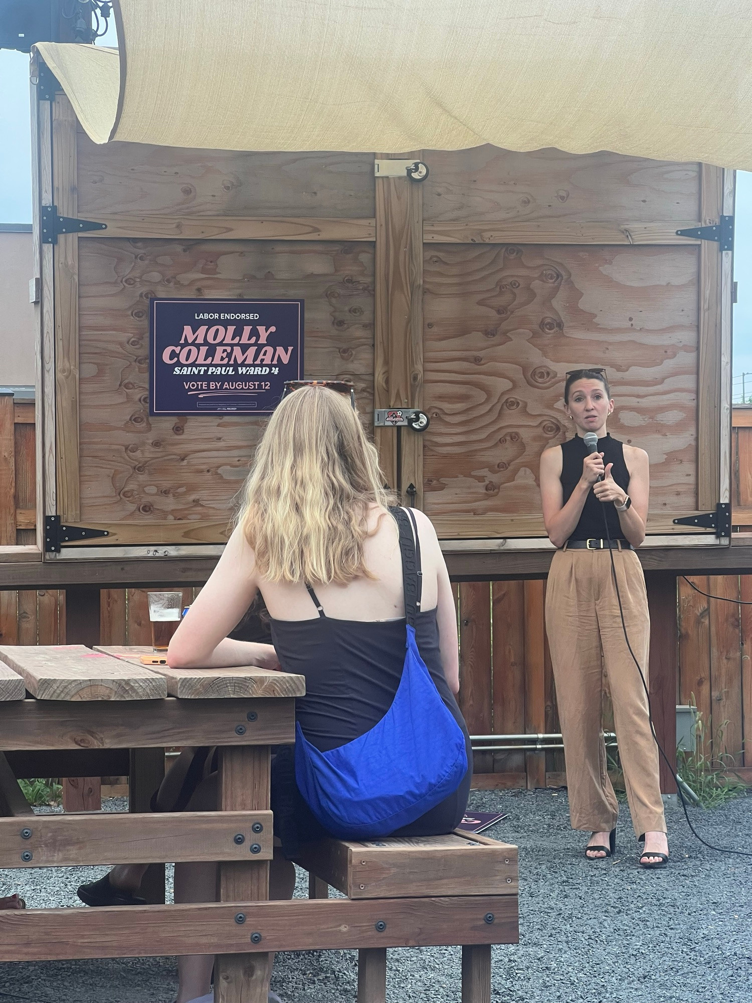 Molly Coleman speaking in on the patio of Black Hart in front of the plywood tv box with a yard sign taped on it. 