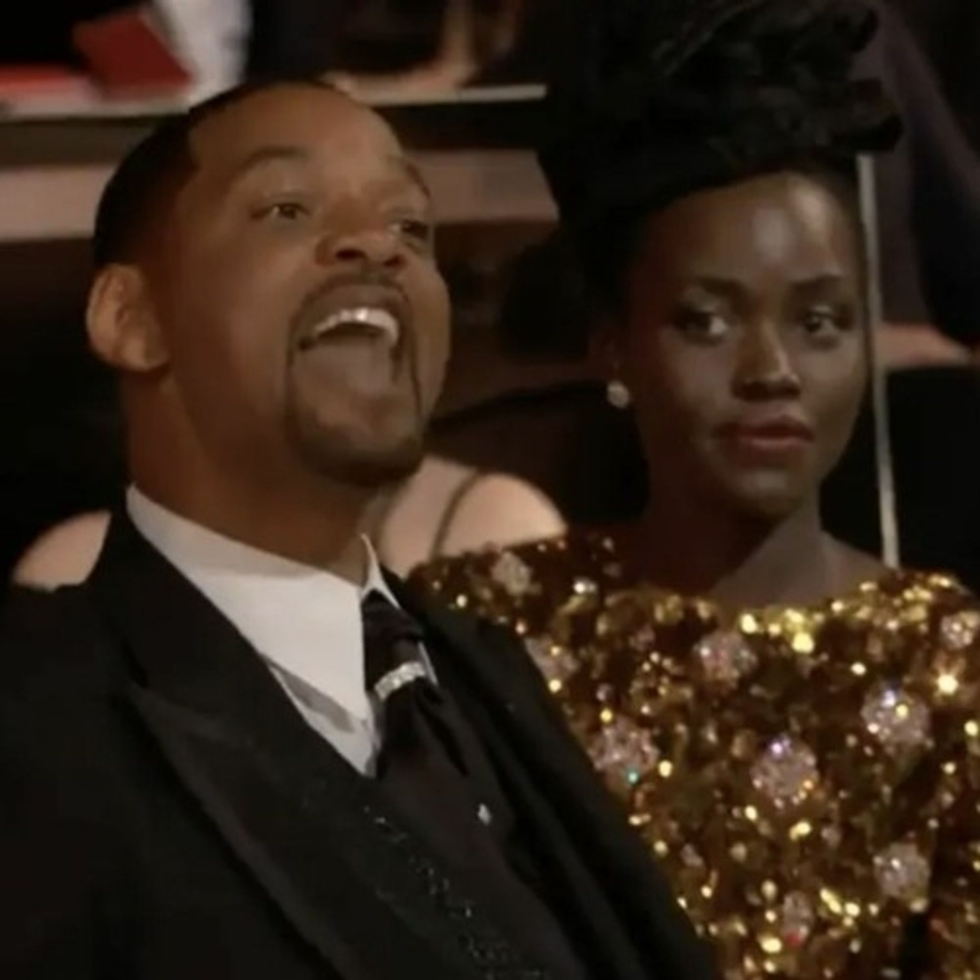 A still photo of Will Smith and his wife at the awards show were he smacked that guy. He looks angry with his mouth wide open. He wears a fancy black suit and tie with white shirt. Jada wears a glittering gold gown next to him.