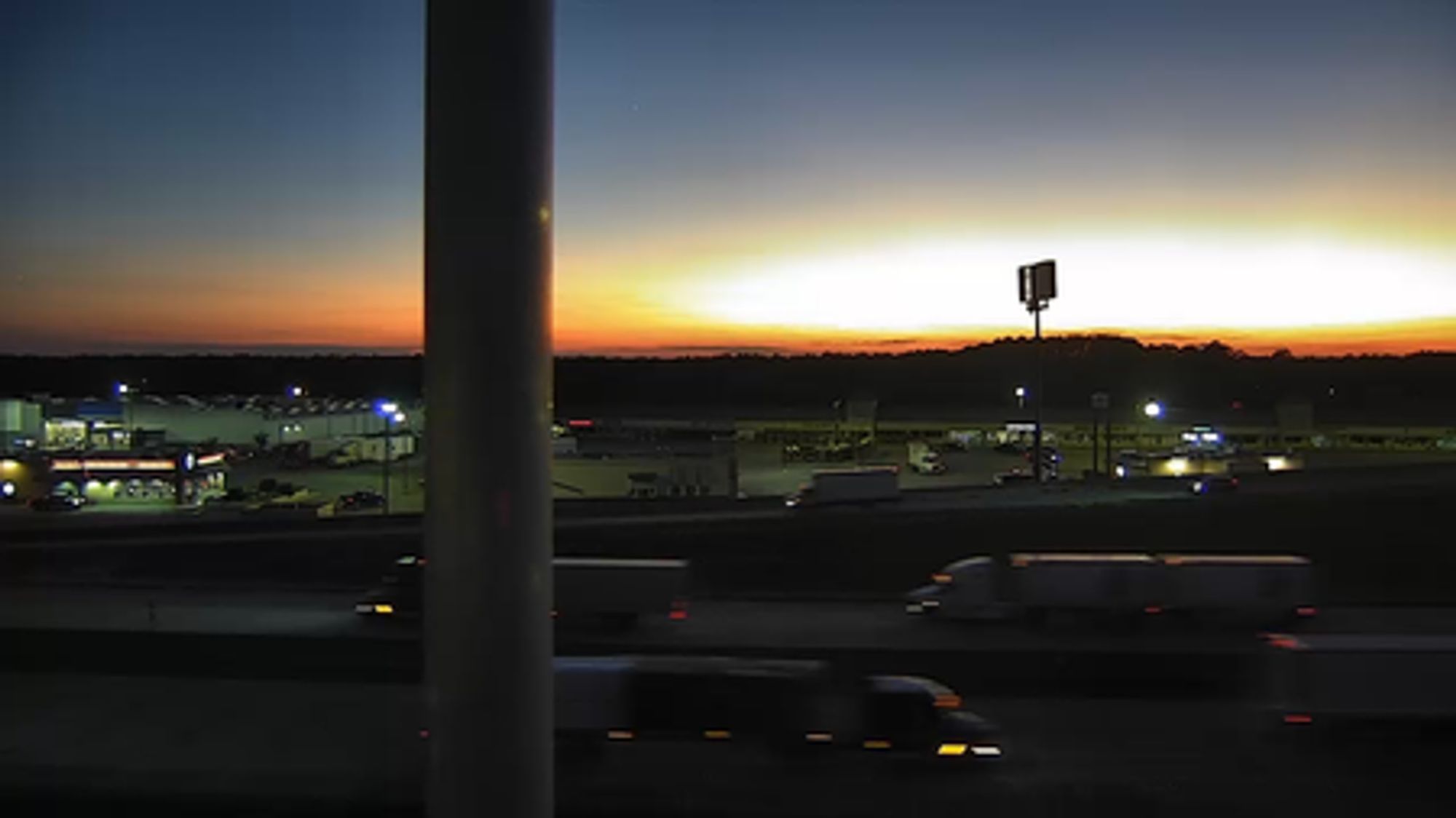 Sunset over a highway, the sun glowing like a nuke on the horizon, a billboard cutting the glow in half.