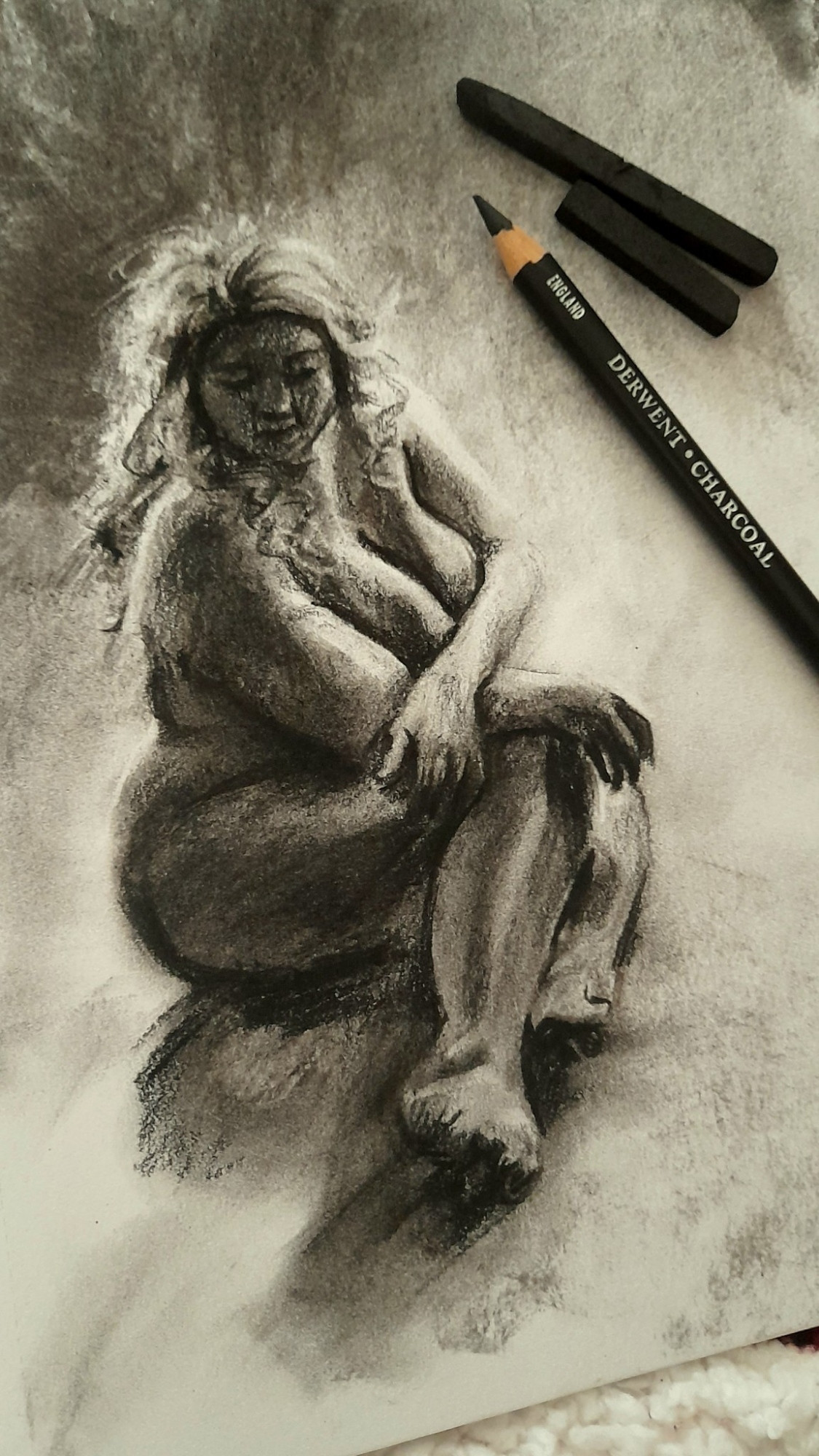 A traditional charcoal sketch of a nude chubby woman with long wavy hair. She brings her knees up to herself, one arm over her large bosom and resting its hand against her other arm, resting against her right leg. 