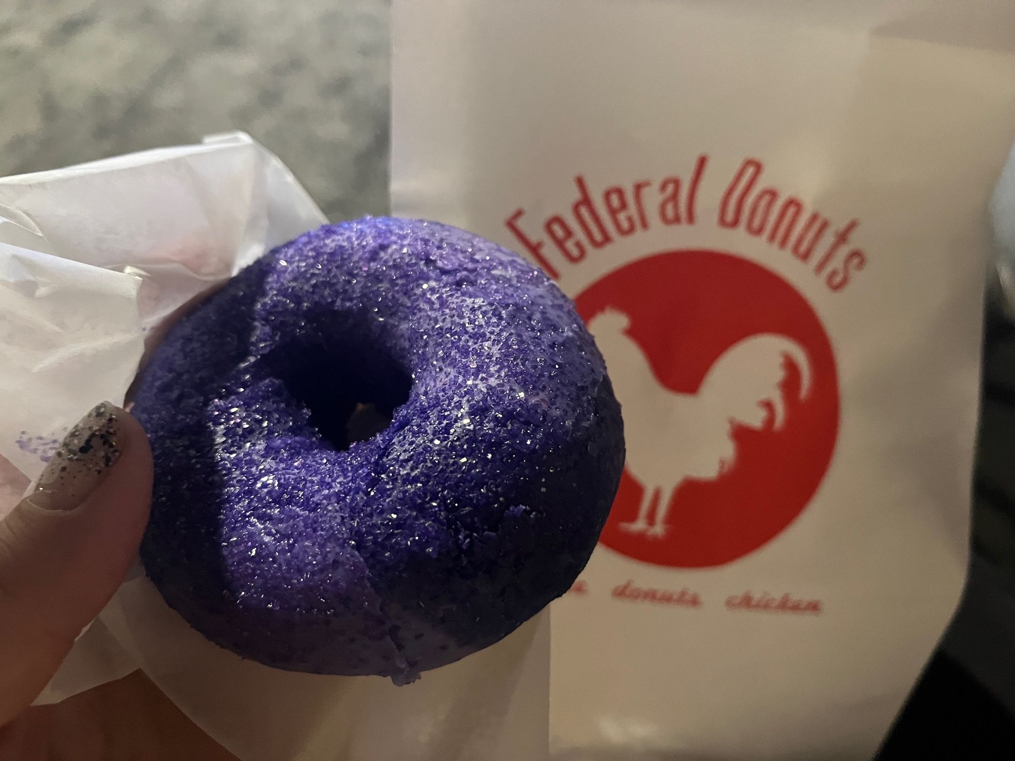 My left hand holding a “Wondrous Space” donut with purply-blue icing and glittery sprinkles. A Federal Donuts paper bag in the background.