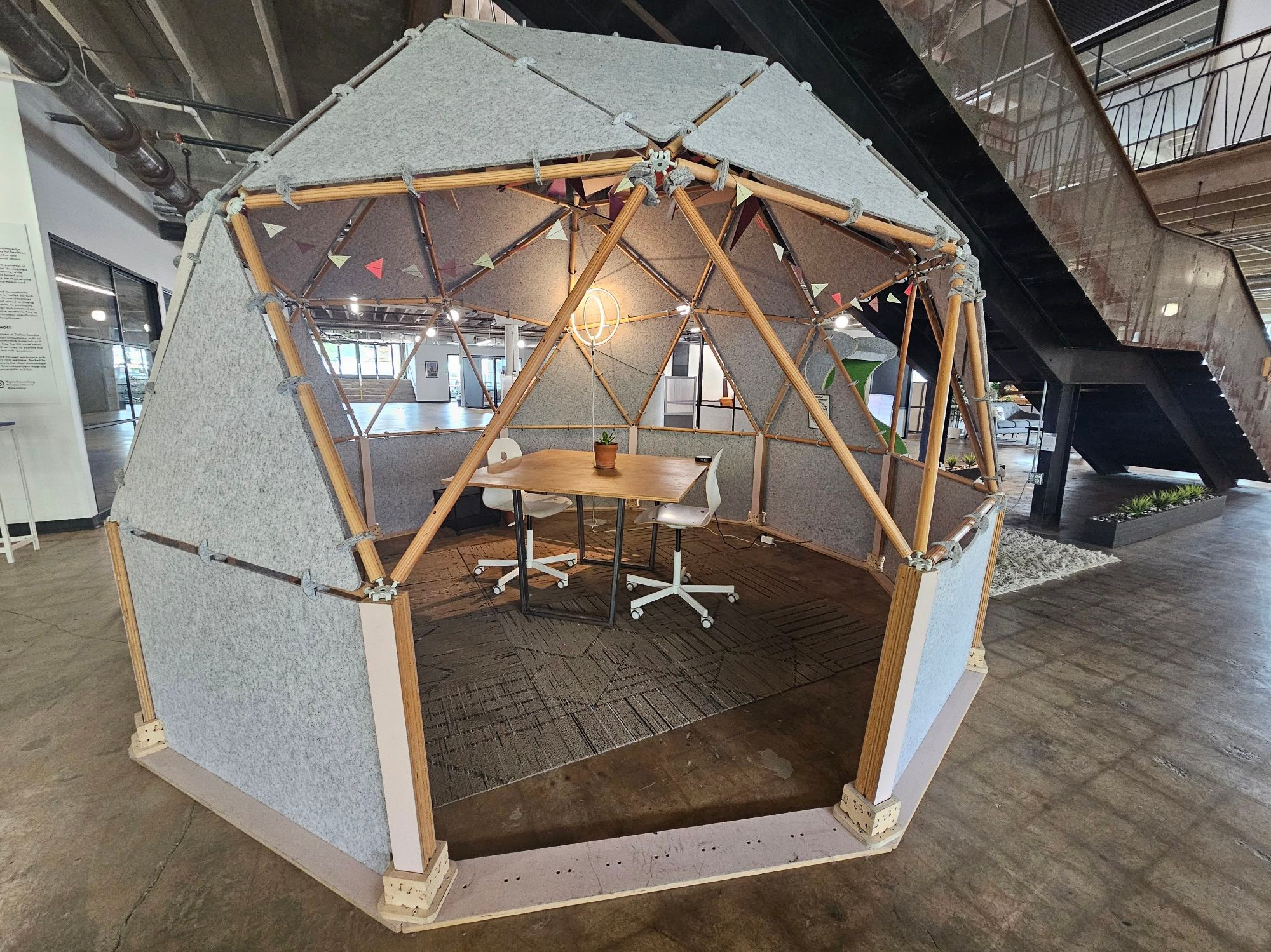 A wood and fabric Geodesic Dome type structure that looks like IKEA made playground inspired office fixtures. Inside are a small table and two chairs, it's set within an industrial building repurposed as an open office floorplan