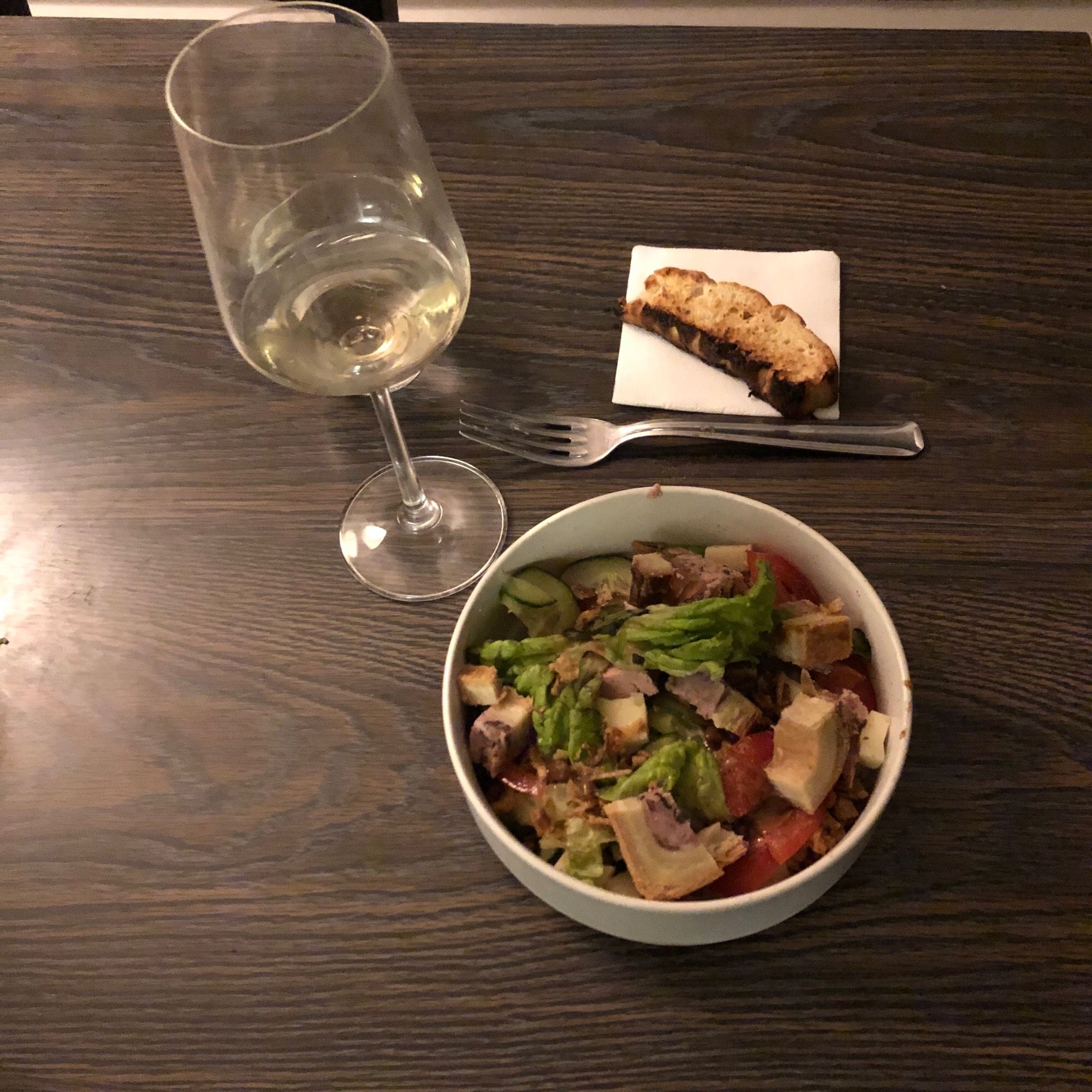 Bol de salade composée avec un verre de vin blanc et un verre d’eau, un bout de pain grillé posé sur une serviette de table à côté d’une fourchette