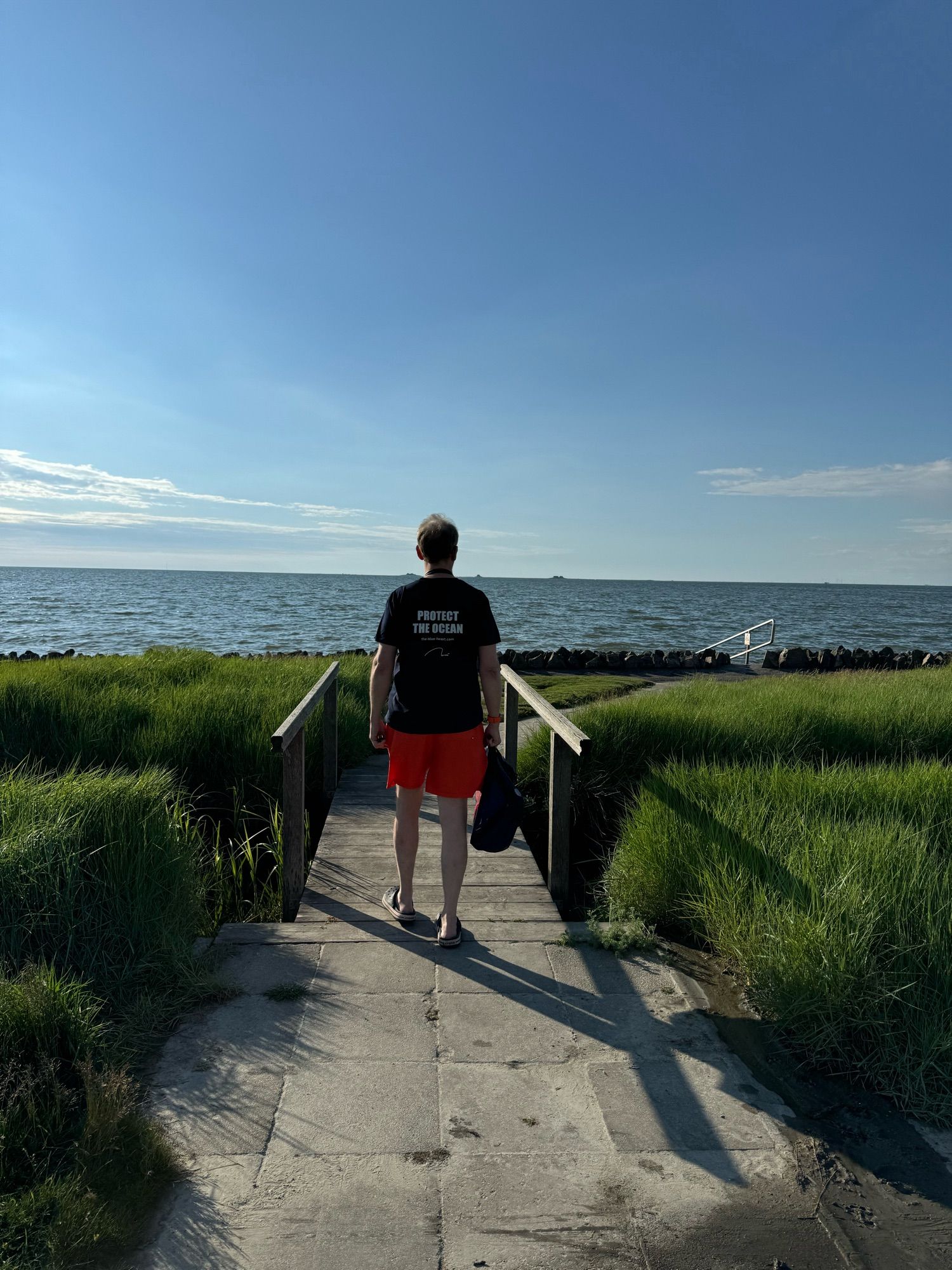 A man walks toward the North Sea under a clear sky. He crosses a small bridge and wear a t-T-shirt with "Protect the Ocean" printed on it.