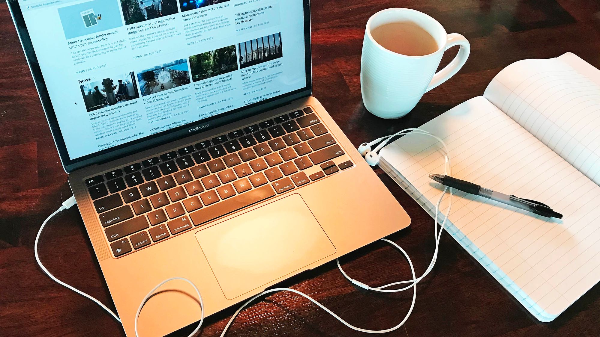 Horizontal photo of a laptop open on a wood grain desk, staged with headphone cords, an open notebook, pen, and mug. Original photo credit Alex Witze.