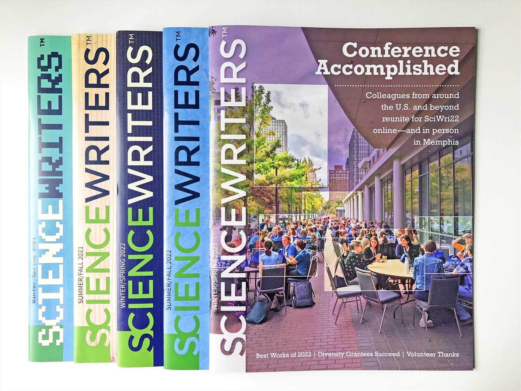 Photograph of the cover of the Winter Spring 2023 Science Writers magazine, laid as the topmost issue across 4 issues of the magazine all lined up together with spine facing to the left, so that the title "Science Writers" repeats neatly in a row.