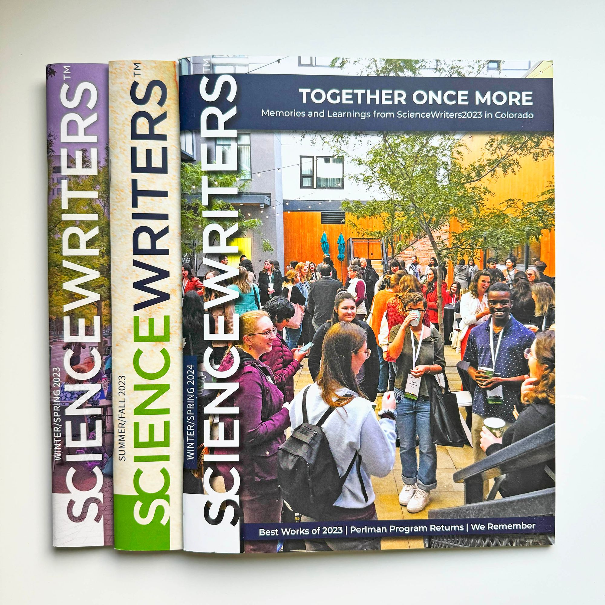 Photograph of the cover of the Winter Spring 2024 Science Writers magazine, laid as the topmost issue across 3 issues of the magazine all lined up together with spine facing to the left, so that the title "Science Writers" repeats neatly in a row.