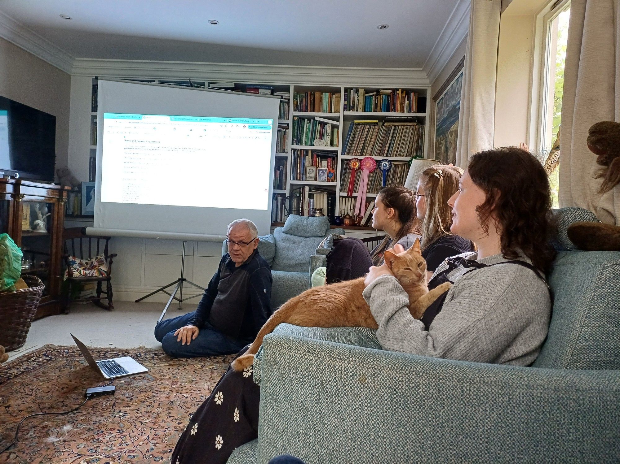 Per sitting on the floor presenting in front of a projector screen that stands in front of a bookcase. On a couch on the right sit Lilia, Jess and Becky with a ginger cat in her lap