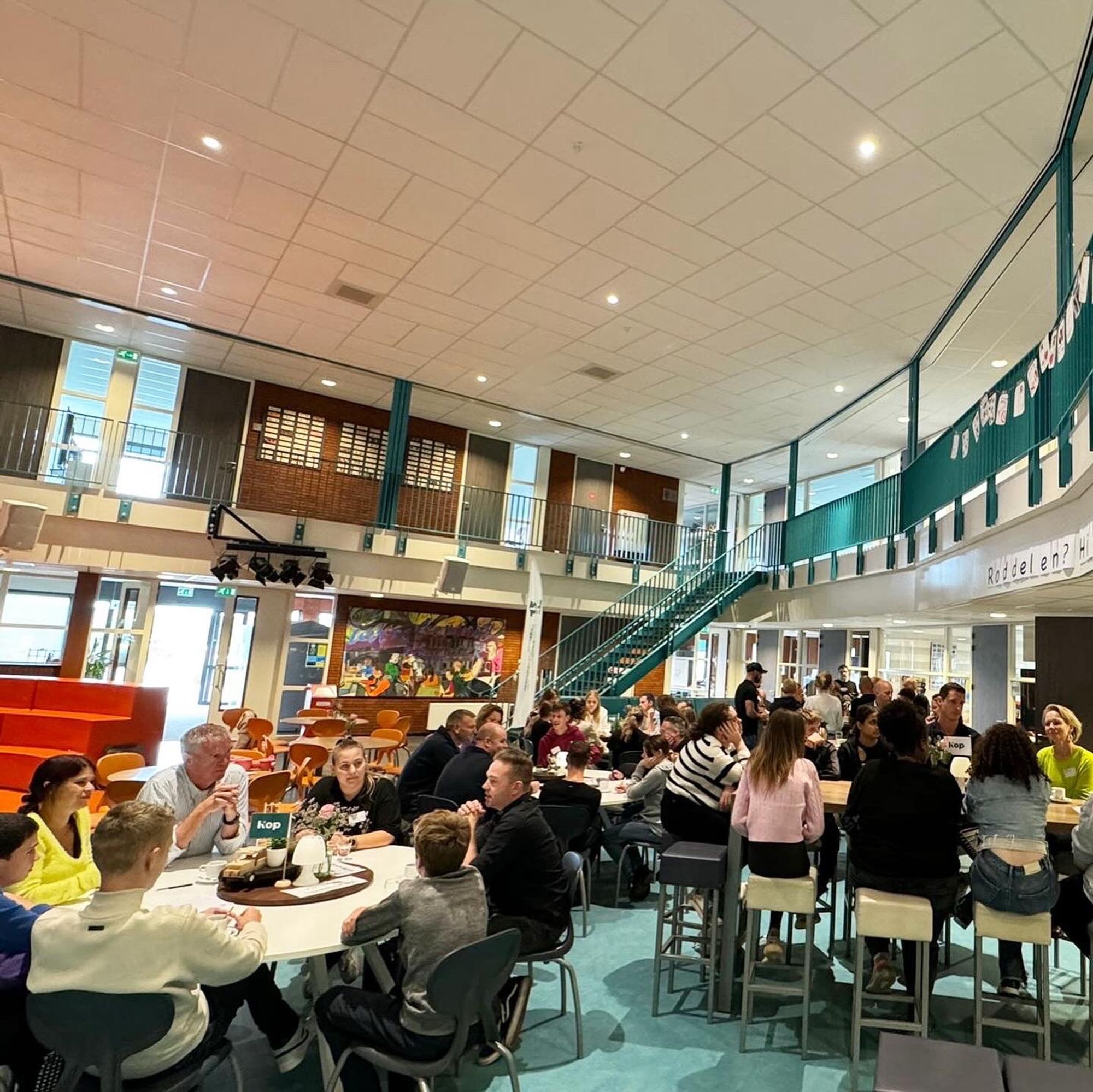 Overzichtsfoto van de aula of centrale plek van dit schoolgebouw. Met verschillende ronde witte tafels waar mensen in groepjes zitten. Op de achtergrond een trap die naar boven gaat, zoals vaker in een schoolgebouw. Met op de eerste verdieping diverse kantoren of lokalen. Er staan ongeveer 40 mensen op deze foto.